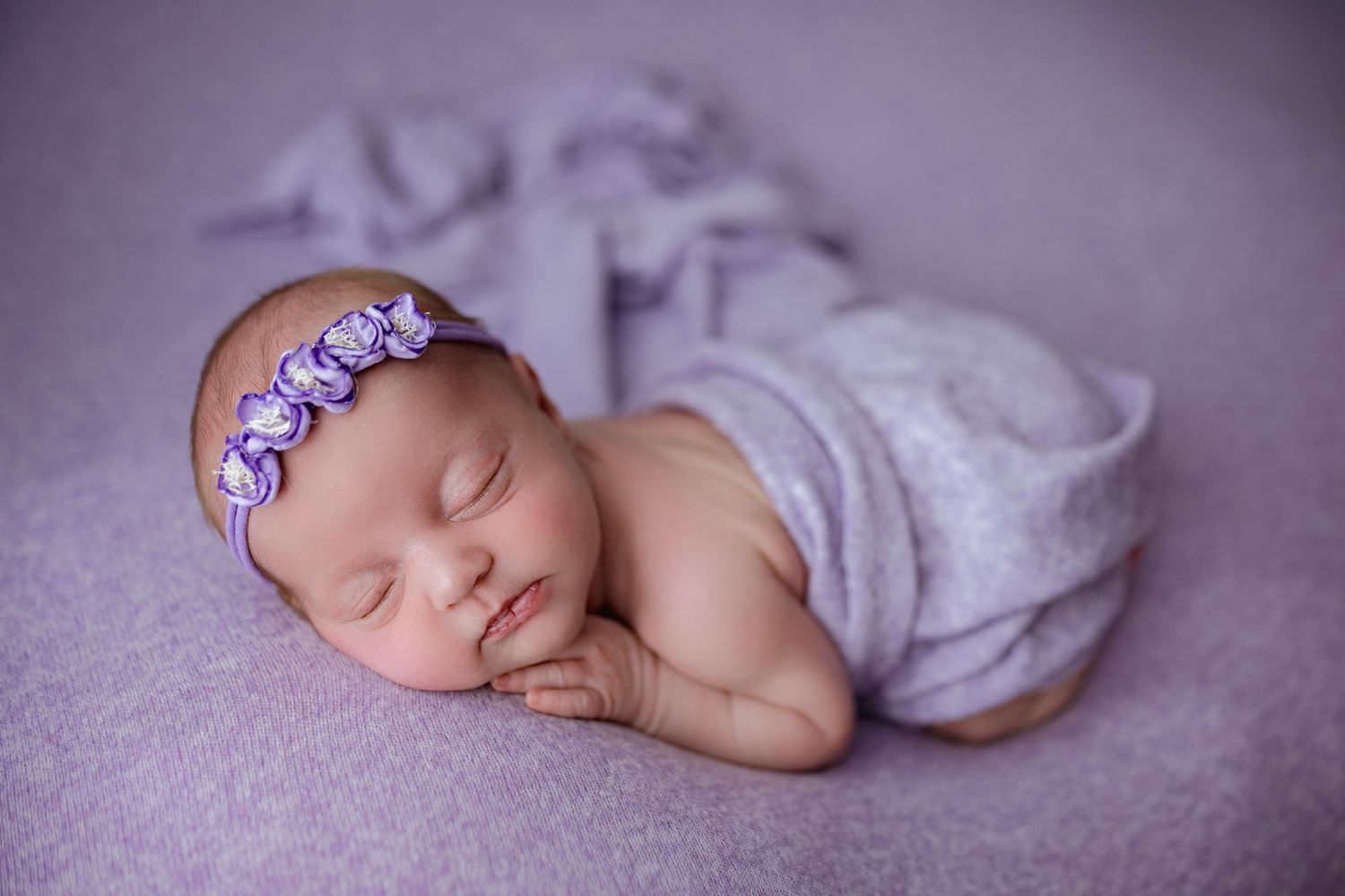 Newborn baby girl sleeping peacefully wrapped in purple cloth with floral headband on lavender background.