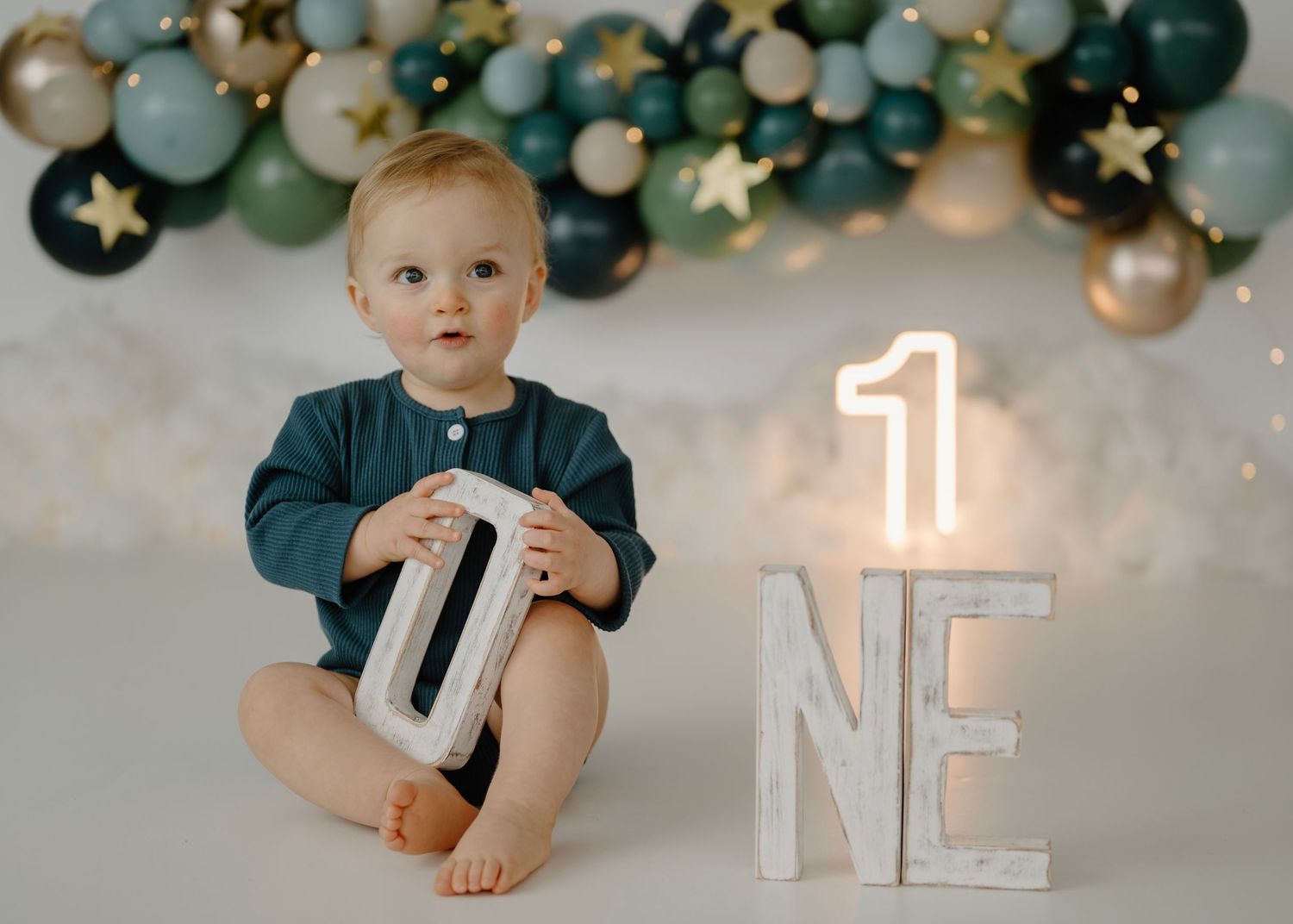 Cute baby boy in teal romper holding ONE letters with star balloon garland backdrop.