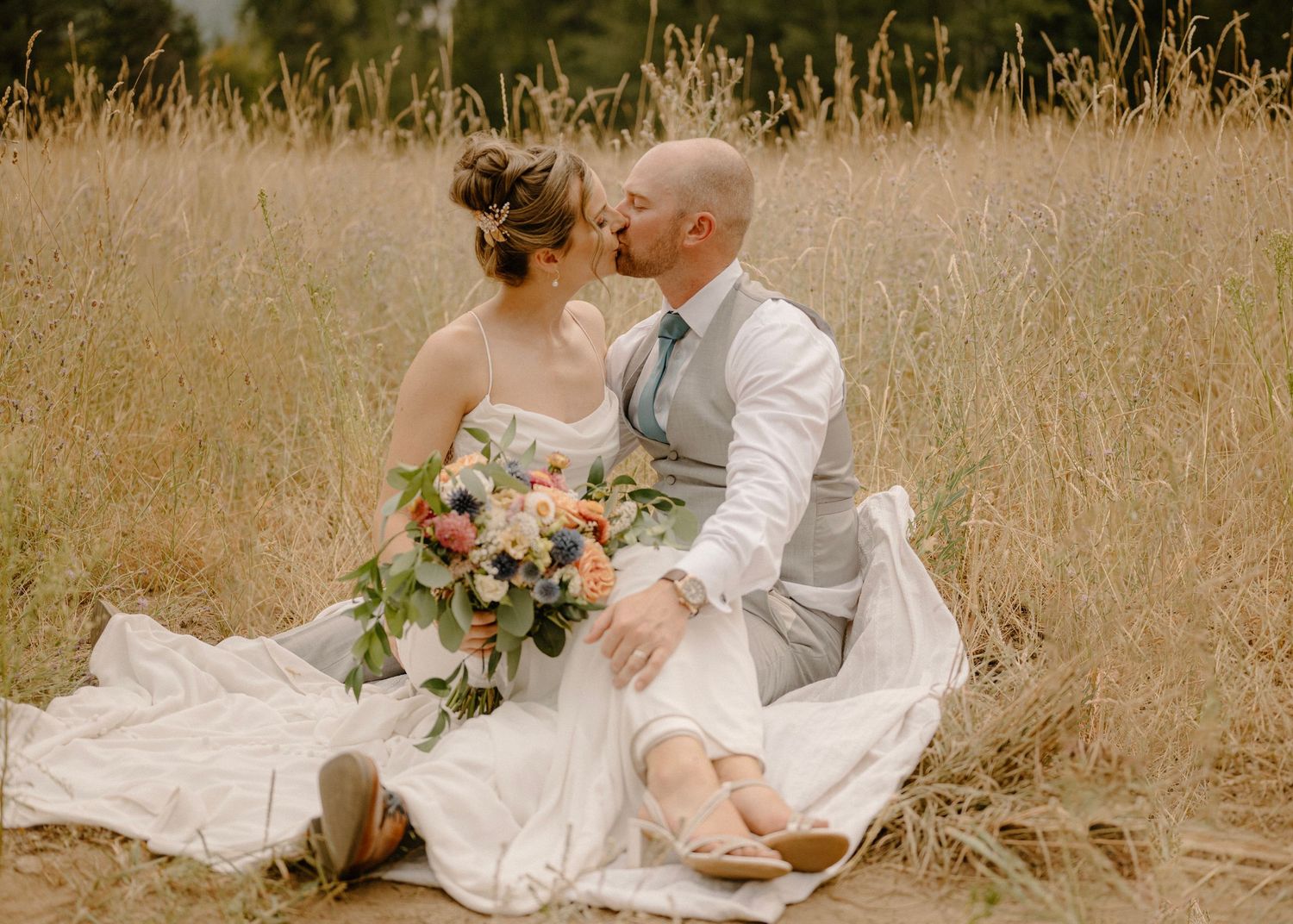 Bride and groom kissing while sitting in a golden wheat field with colorful bridal bouquet.