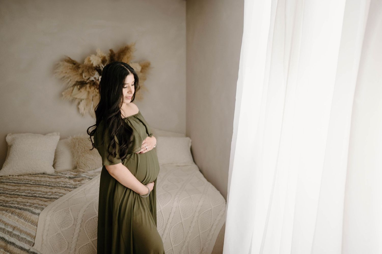 Pregnant woman in olive off-shoulder gown standing between bed and sheer white curtains.