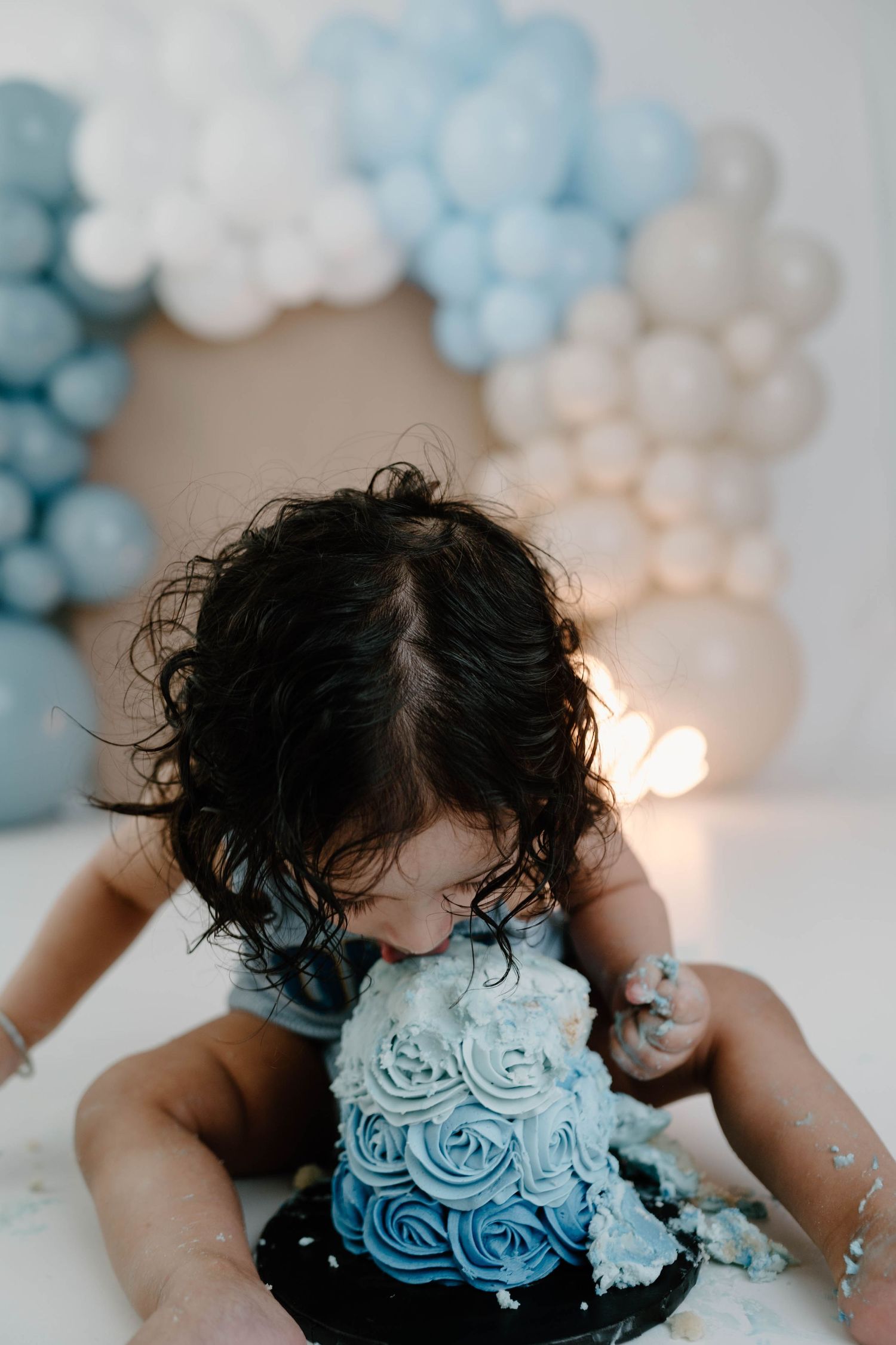 A toddler with curly dark hair leans face-first into a blue ombre rosette cake with balloon decorations behind.