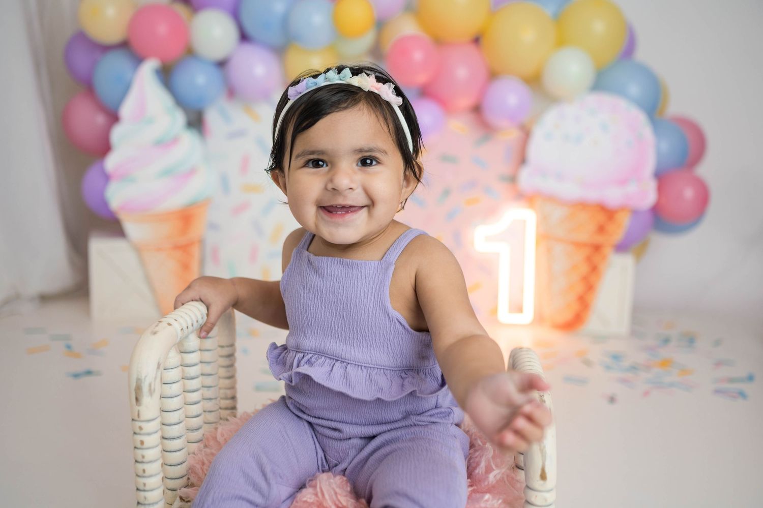 Happy baby girl in purple outfit sits in wicker chair at colorful ice cream first birthday celebration in Leduc.
