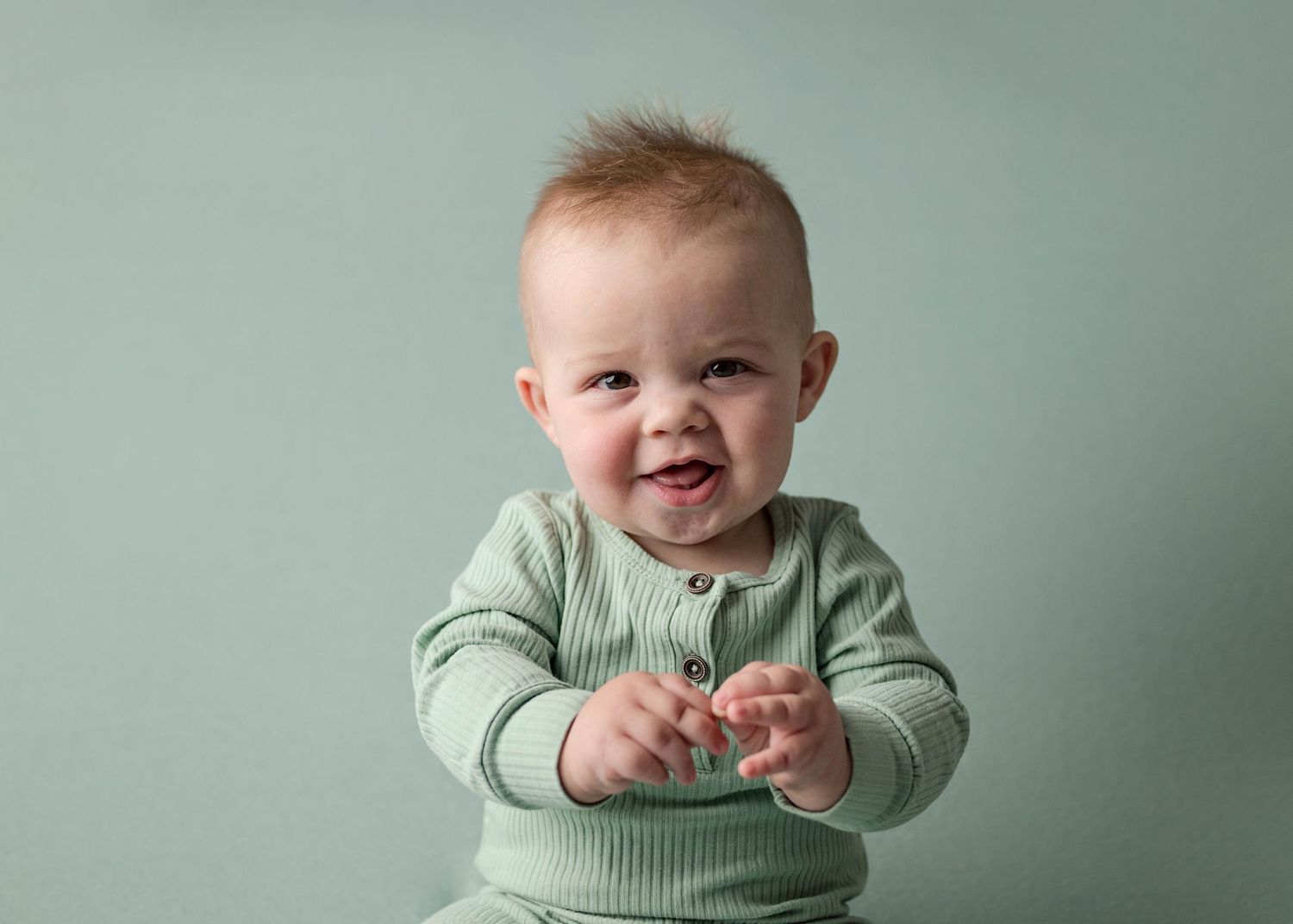 Smiling red-haired baby in sage green ribbed romper sitting and clapping hands during studio photo session.