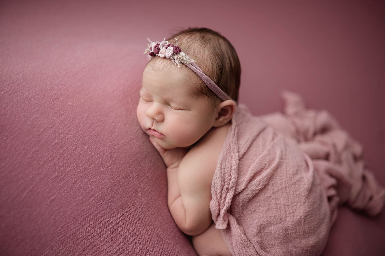 Sleeping newborn baby girl wrapped in pink with floral headband on a soft pink background.