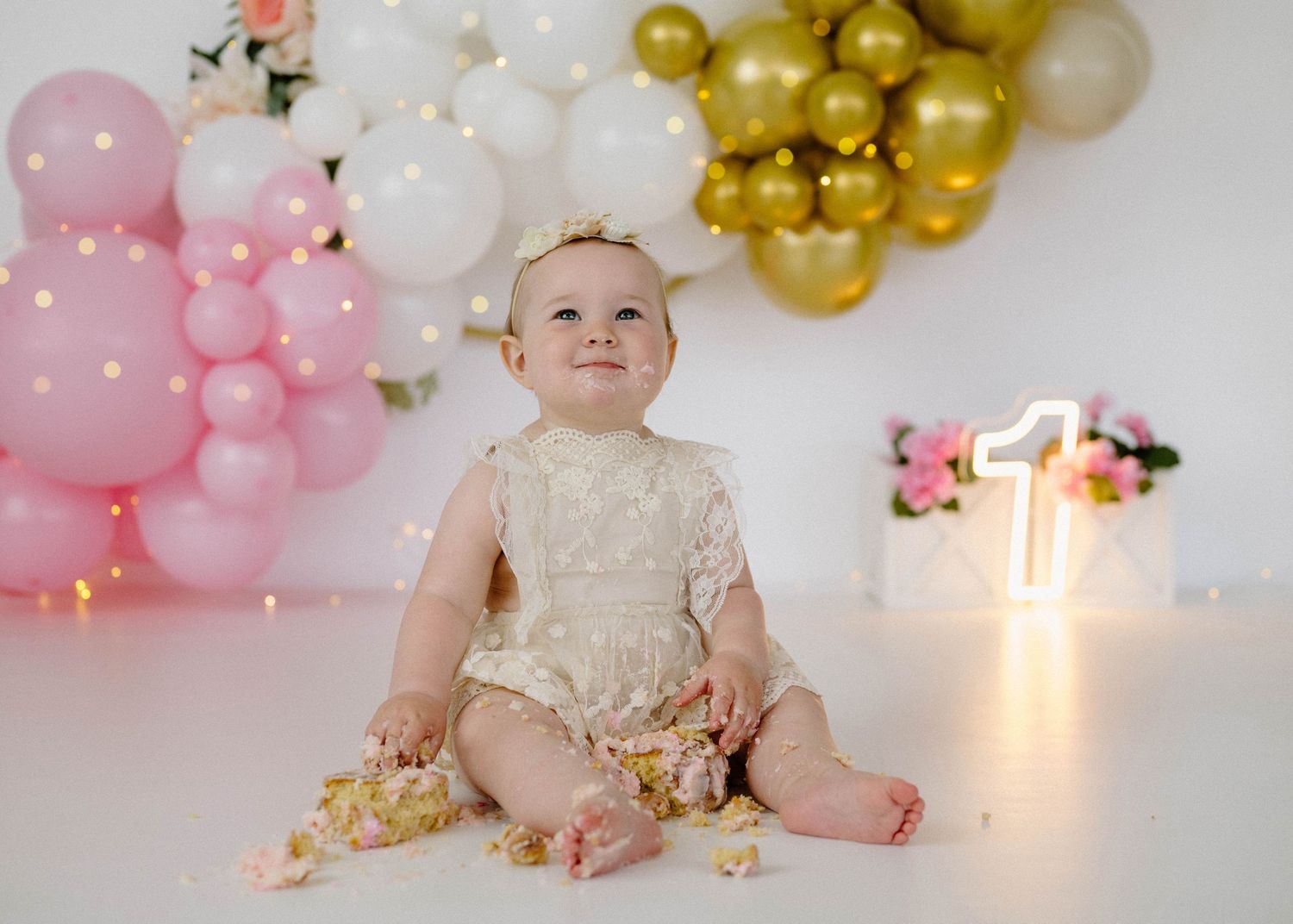 Baby girl in messy lace romper sits on floor after cake smash at first birthday with balloon garland.