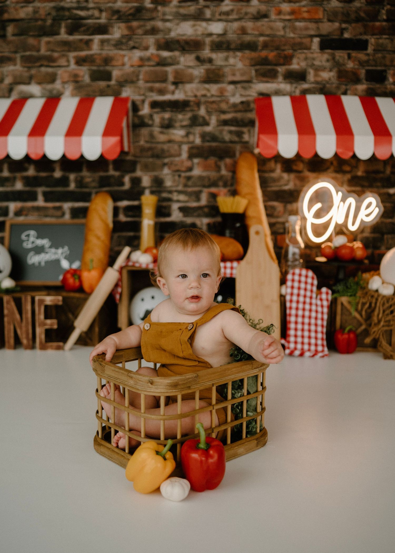 Baby in rust overalls sits in wooden crate surrounded by vegetables at Italian-themed birthday shoot.