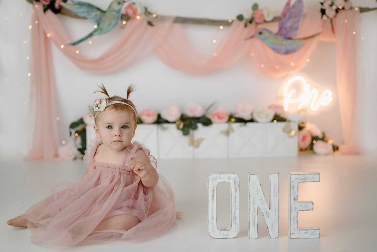 Baby girl in pink dress sits beside illuminated ONE letters at a hummingbird floral first birthday session.