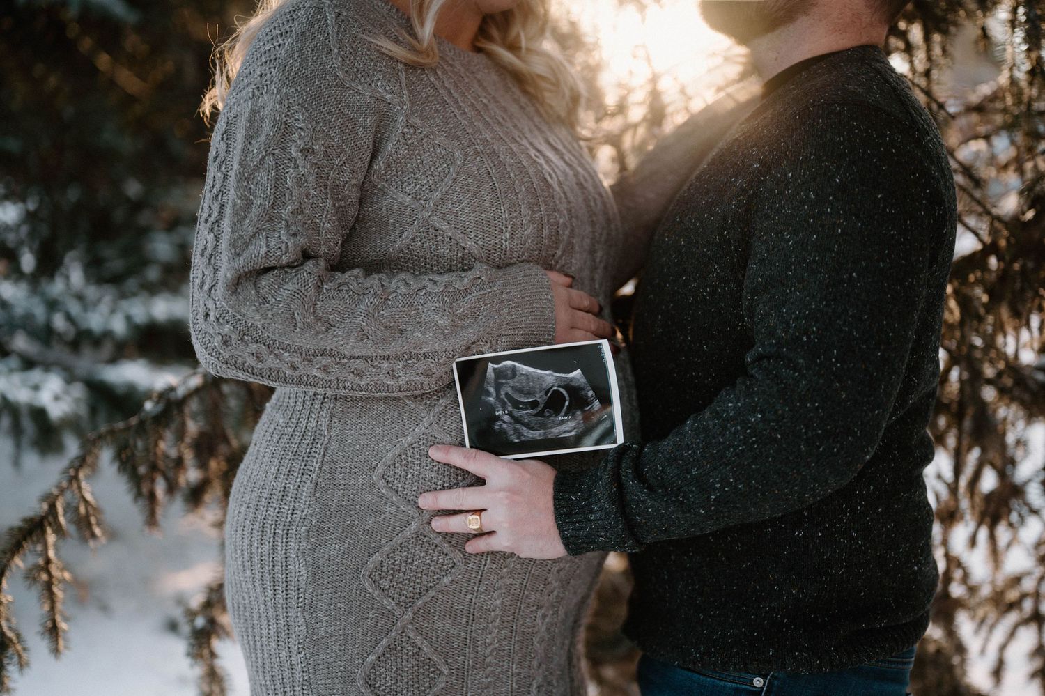 Couple holding ultrasound photo against pregnant belly under snowy pine trees at sunset in Fred Johns Park in Leduc.
