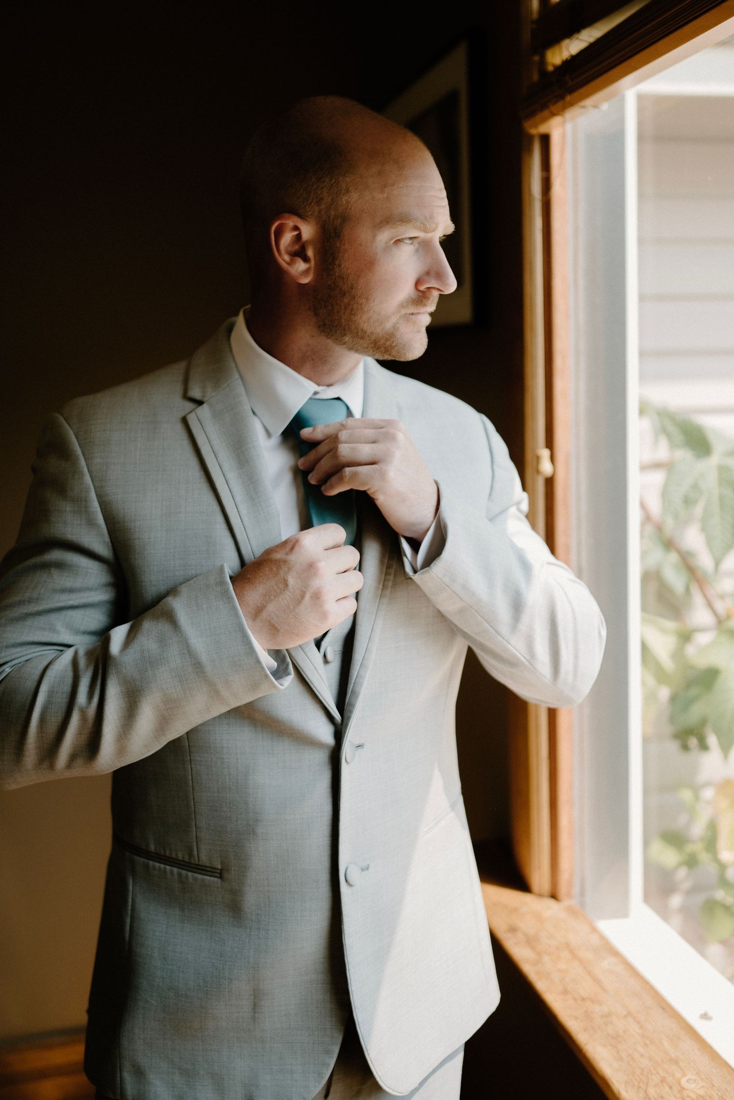 Bald groom in grey suit adjusting teal tie while standing by a sunlit window before wedding.