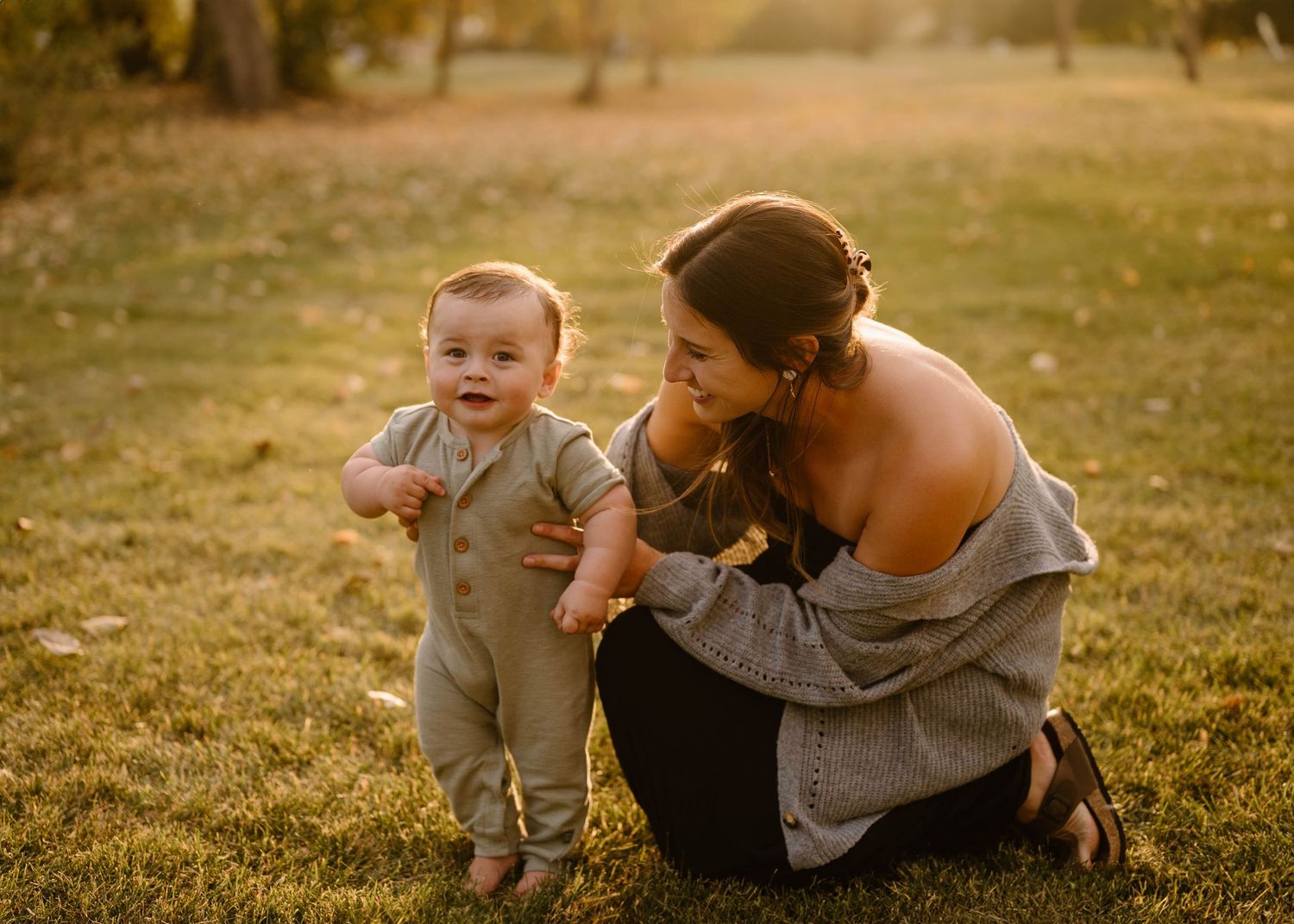 Mother kneeling on grass helping her baby boy take first steps in sunny autumn park.