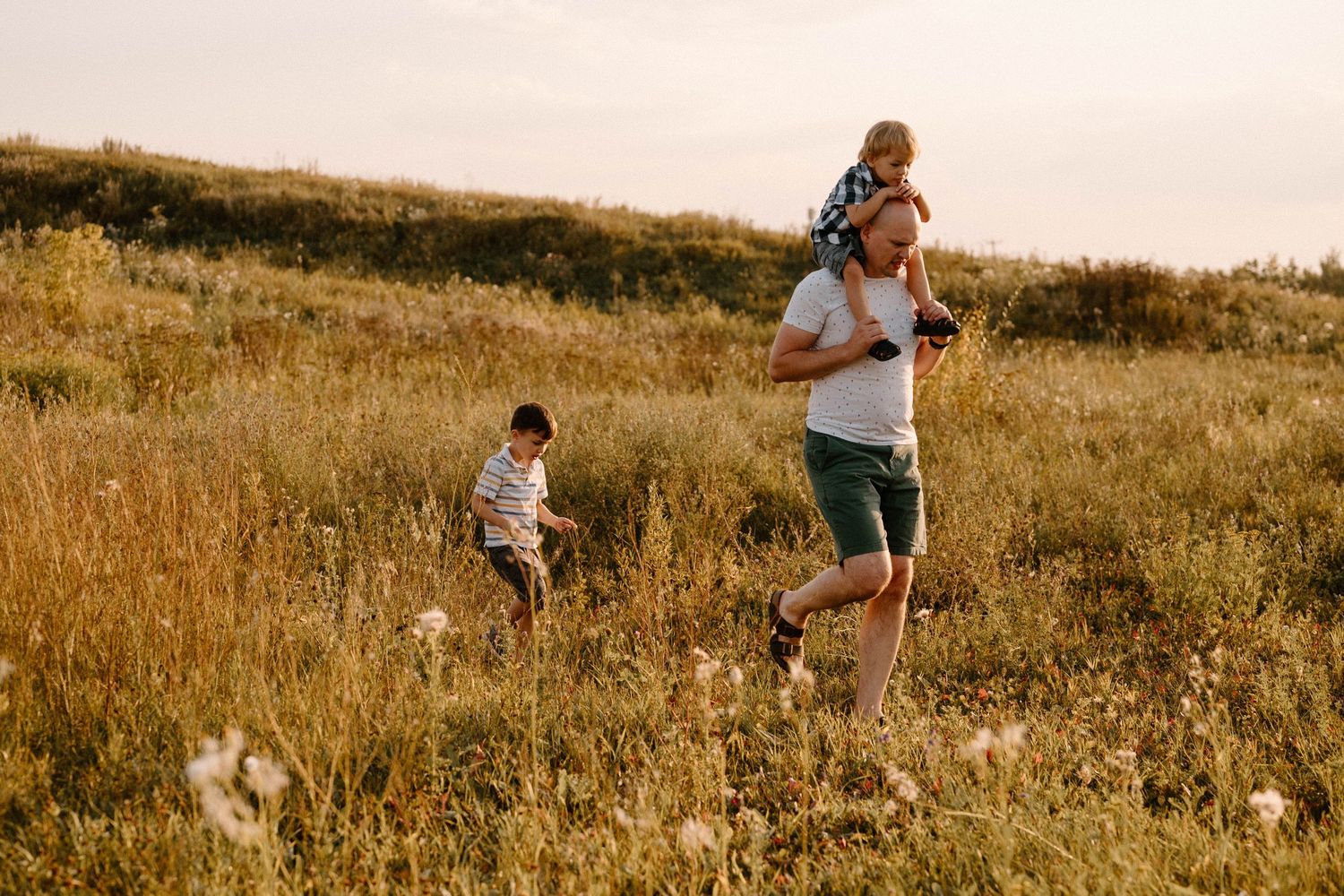 Father carrying child on shoulders while another child runs through a sunny meadow.