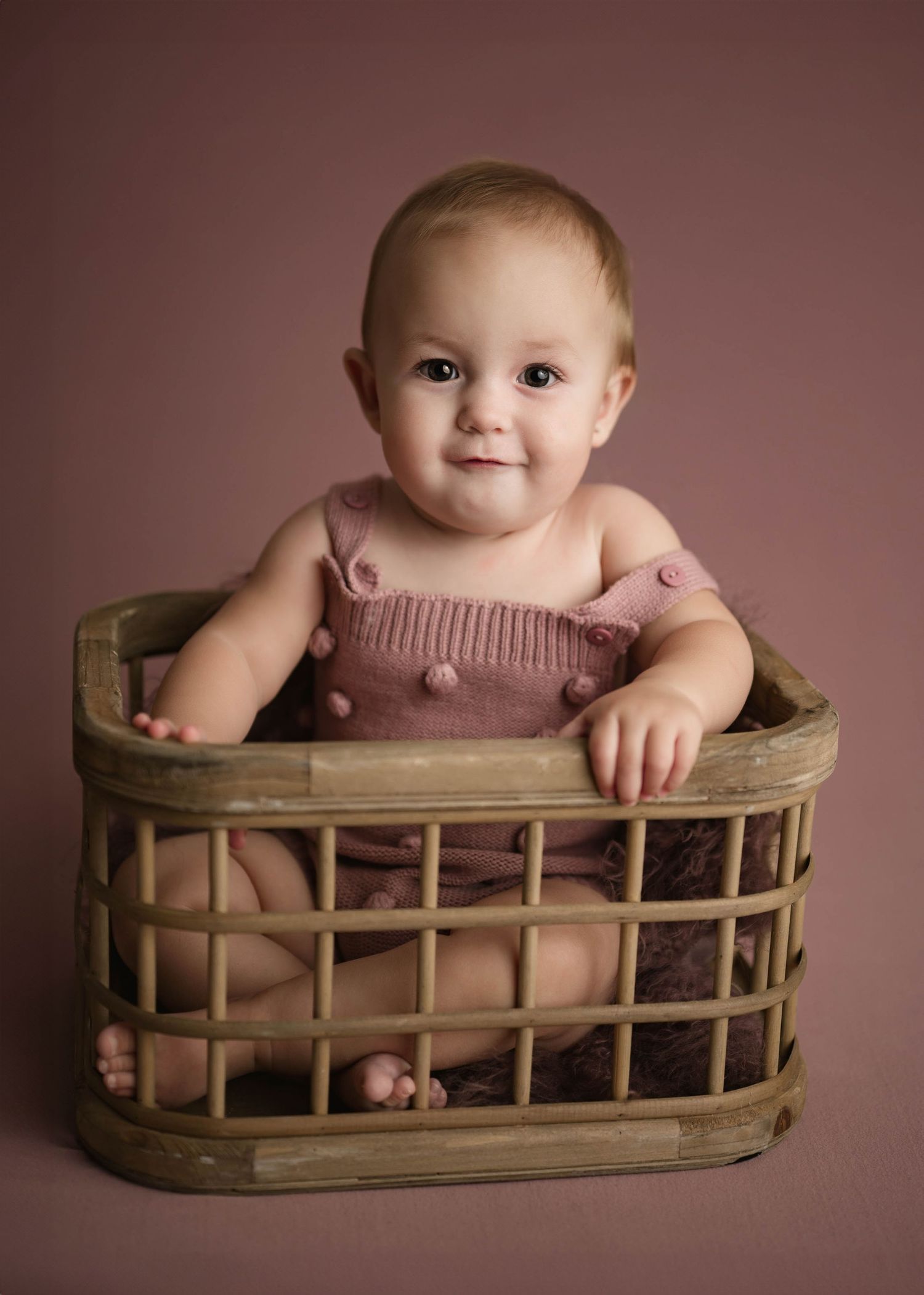 Cute baby girl in pink knit romper sitting inside rustic wooden basket on pink background.
