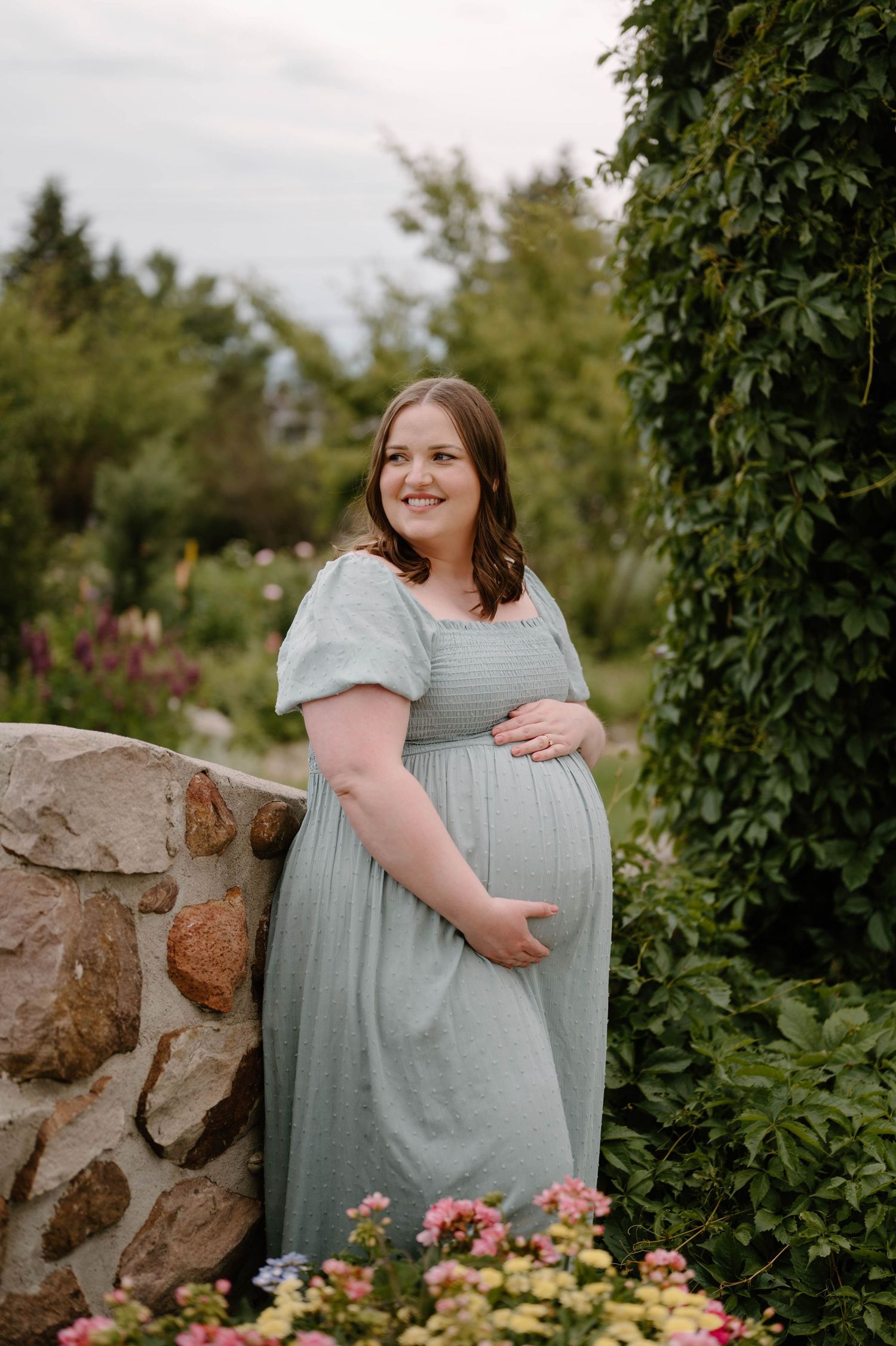 Pregnant woman in sage green dress smiling in a lush garden with stone wall and colorful flowers.