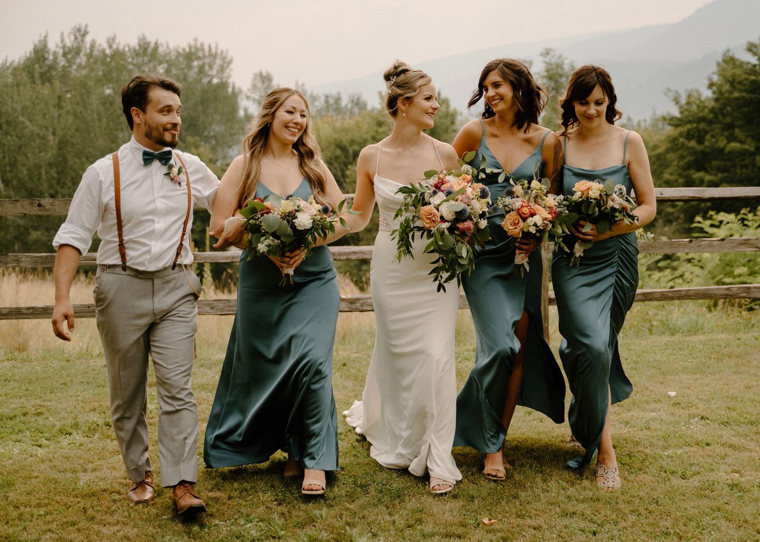 Bride with bridesmaids in teal dresses walking and laughing together outdoors with lush greenery behind them.
