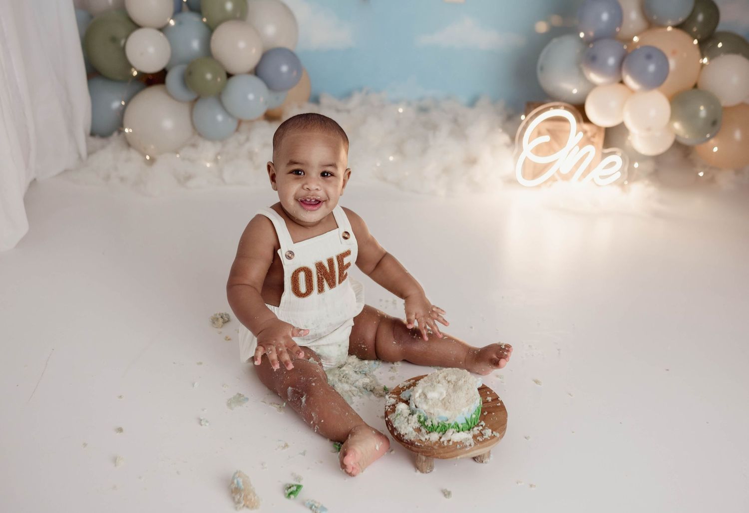 Smiling one-year-old boy sitting with smashed birthday cake, wearing ONE outfit near neon sign.