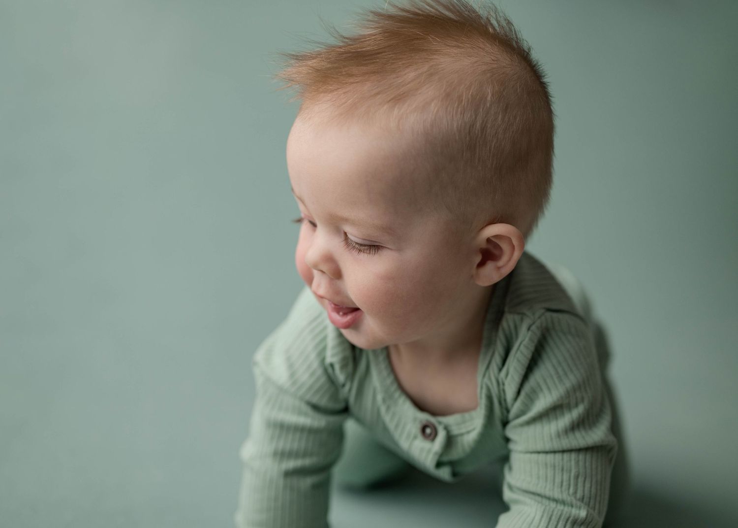 Side profile of red-haired baby in mint green romper sitting up during professional milestone photo session.