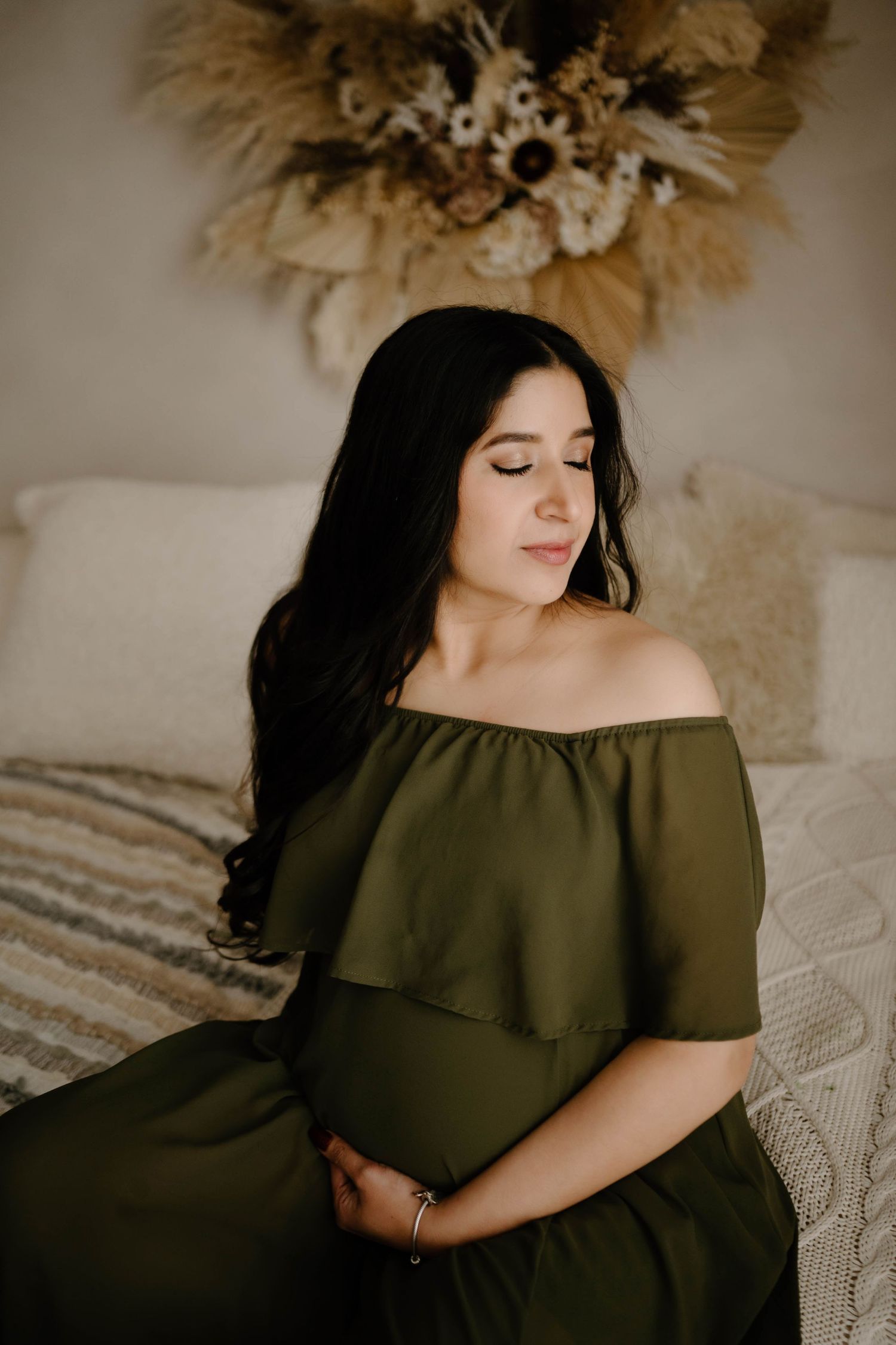 Pregnant woman in olive off-shoulder dress seated on bed with boho pampas decor in a Leduc photo studio.