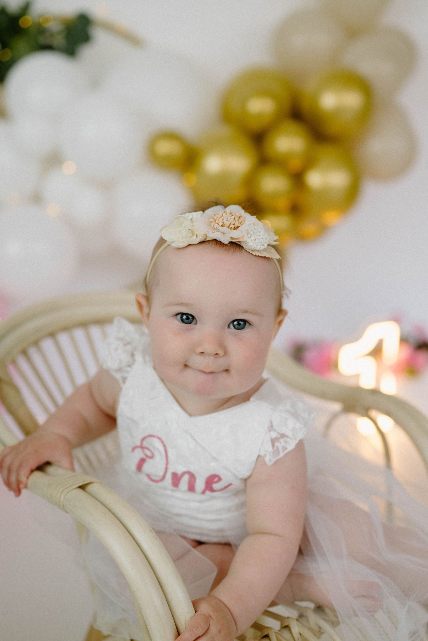 Close-up of baby girl in white One romper with floral headband sitting in rattan chair at first birthday.