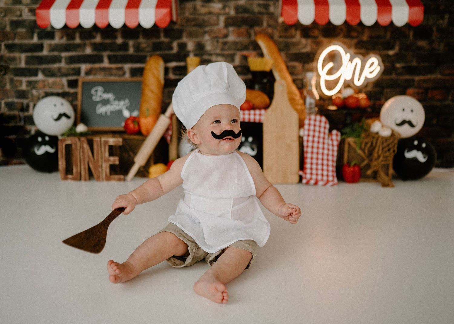 Baby in chef hat holds wooden spoon with fake mustache at Italian-themed first birthday cake smash.