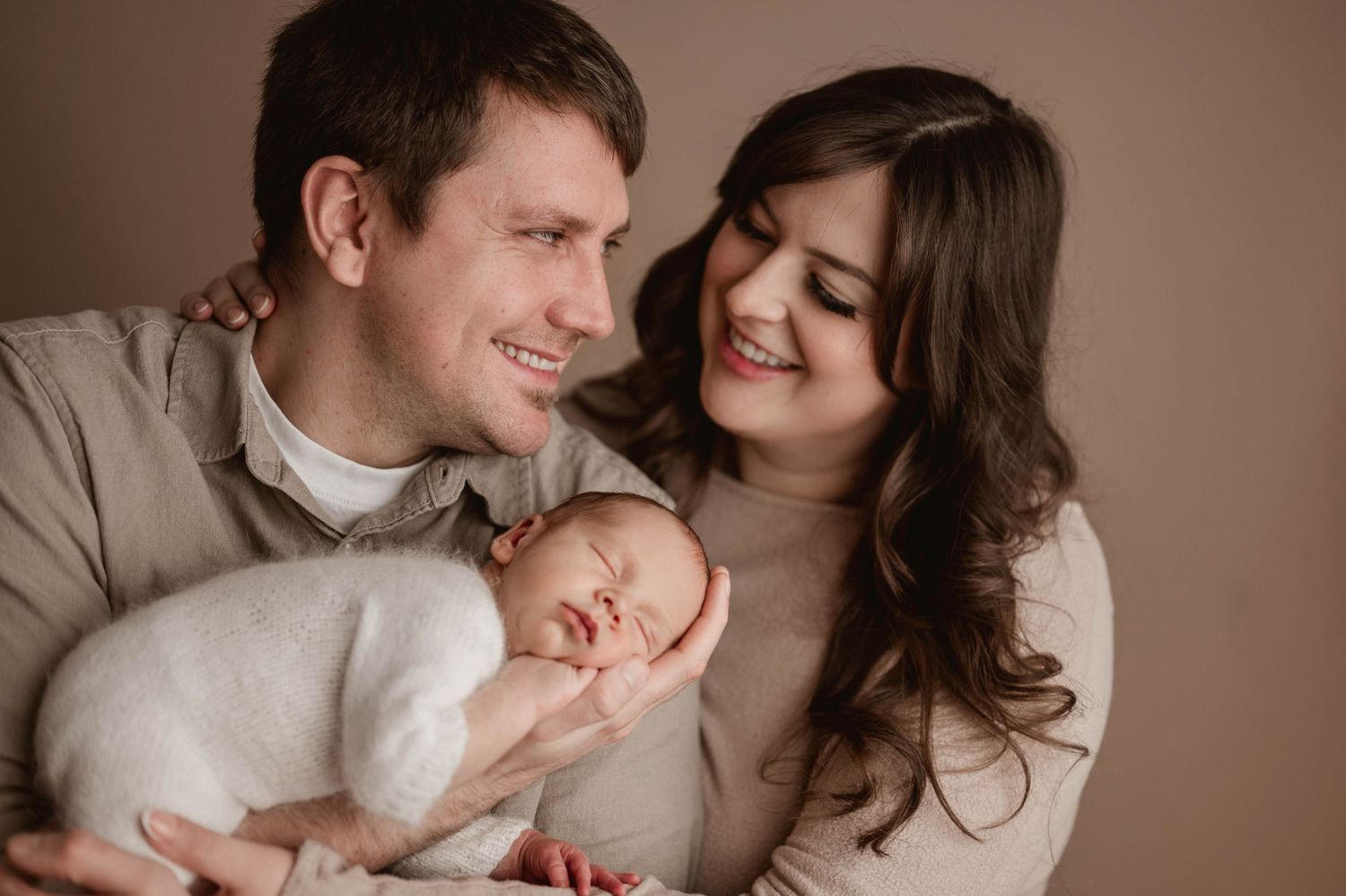 Smiling parents holding their sleeping newborn baby wrapped in white during a studio portrait.