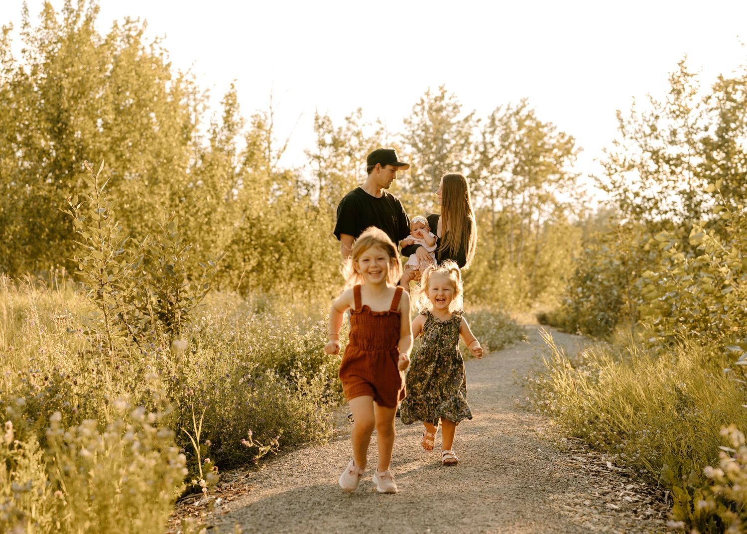 Father walking behind two young daughters on a sunlit path through lush greenery.
