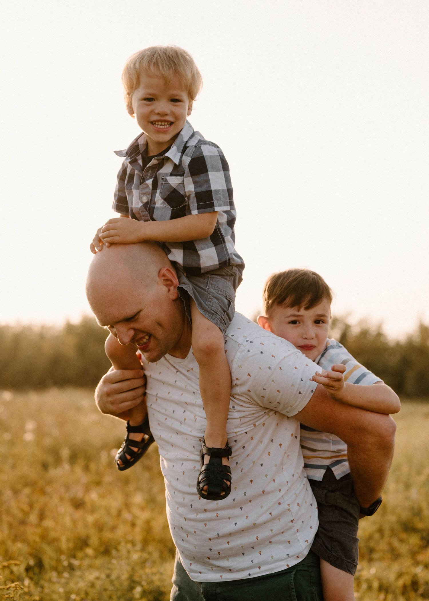 Smiling bald father carrying two young boys on shoulders and arms in sunny field.