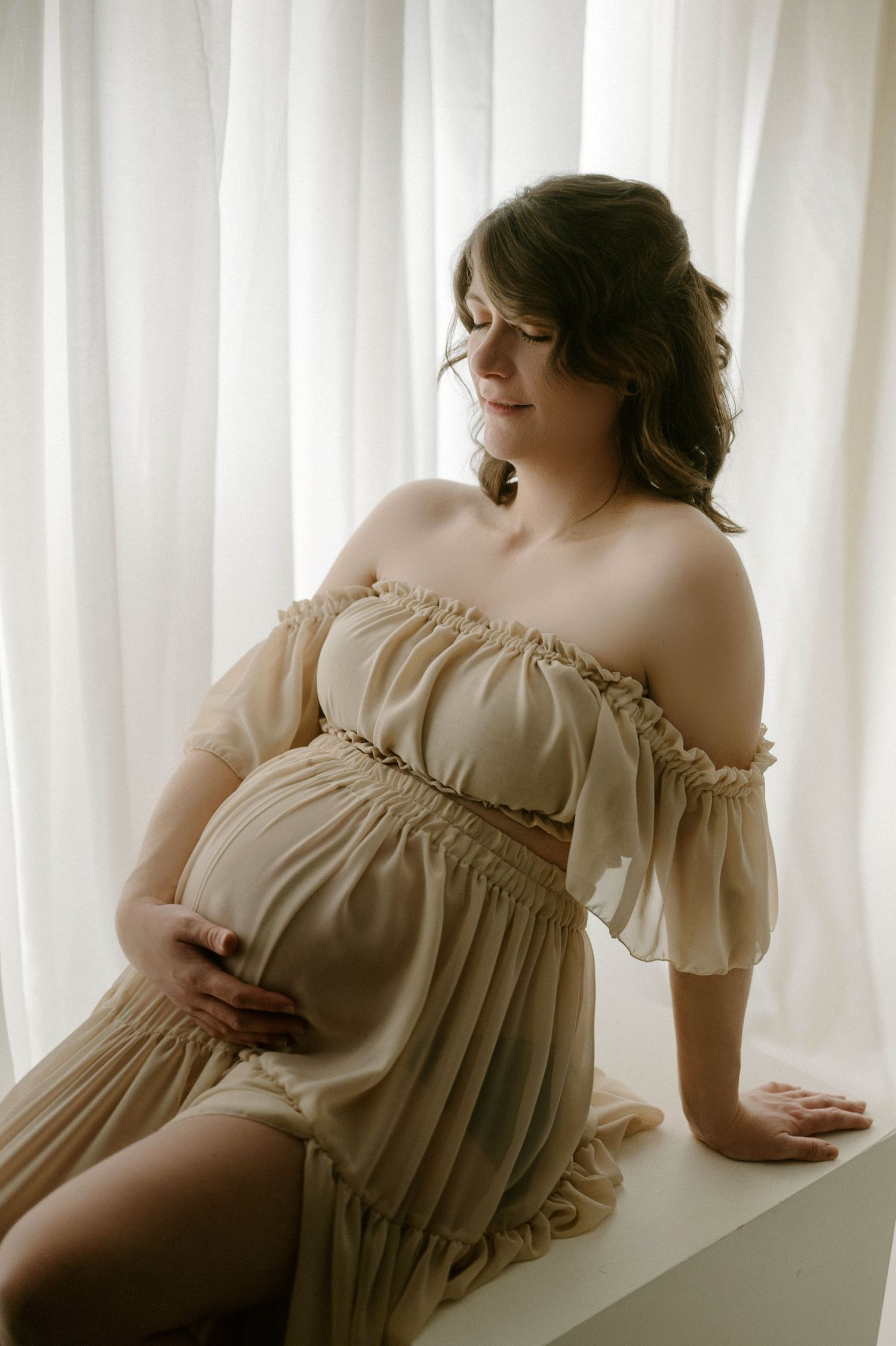 Maternity photo of pregnant woman in off-shoulder beige dress gently holding baby bump near white curtains.