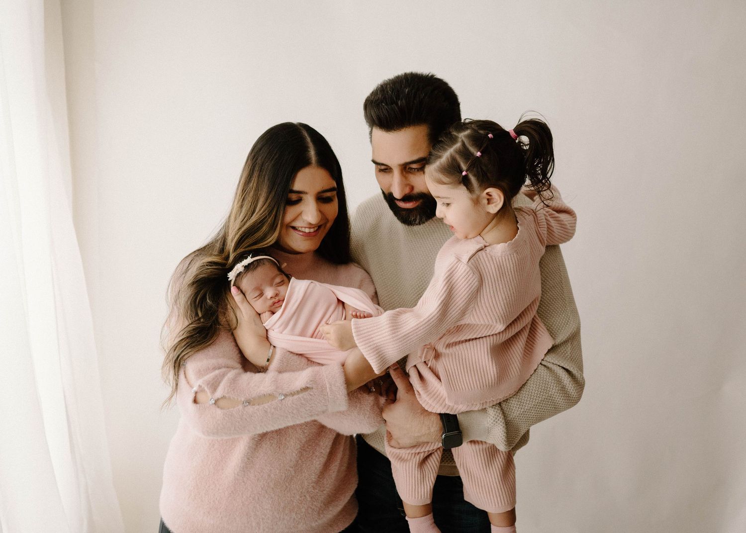 Family portrait with parents in pink holding newborn and toddler daughter against white studio background.
