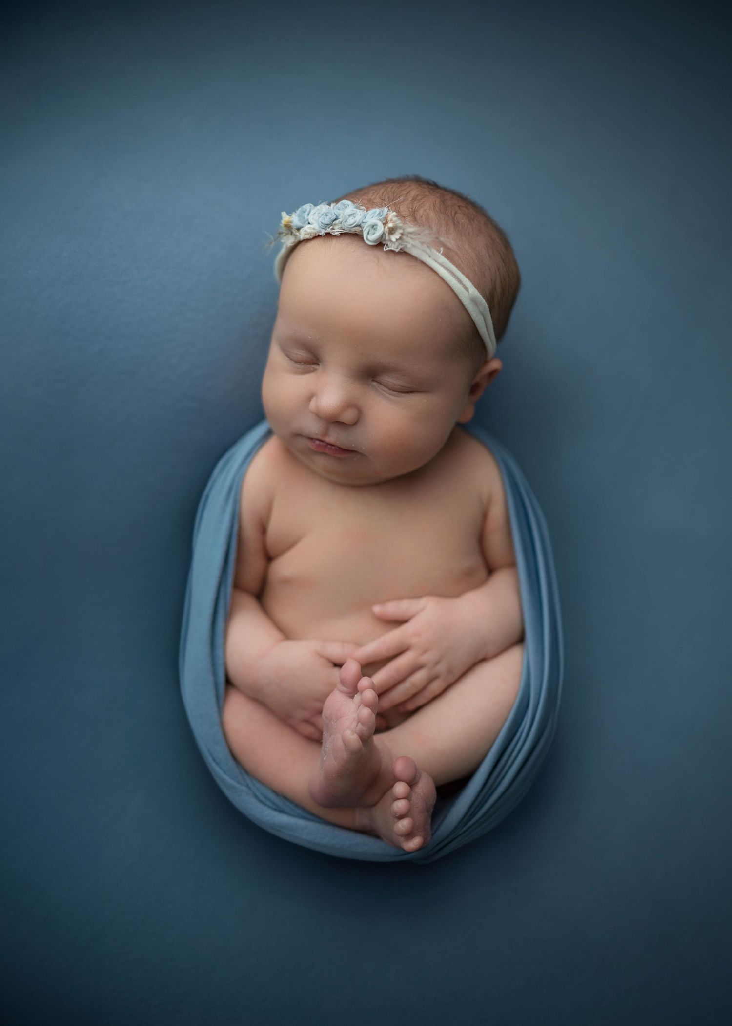 Newborn baby girl wrapped in blue fabric with a floral headband, peacefully sleeping on a blue background.