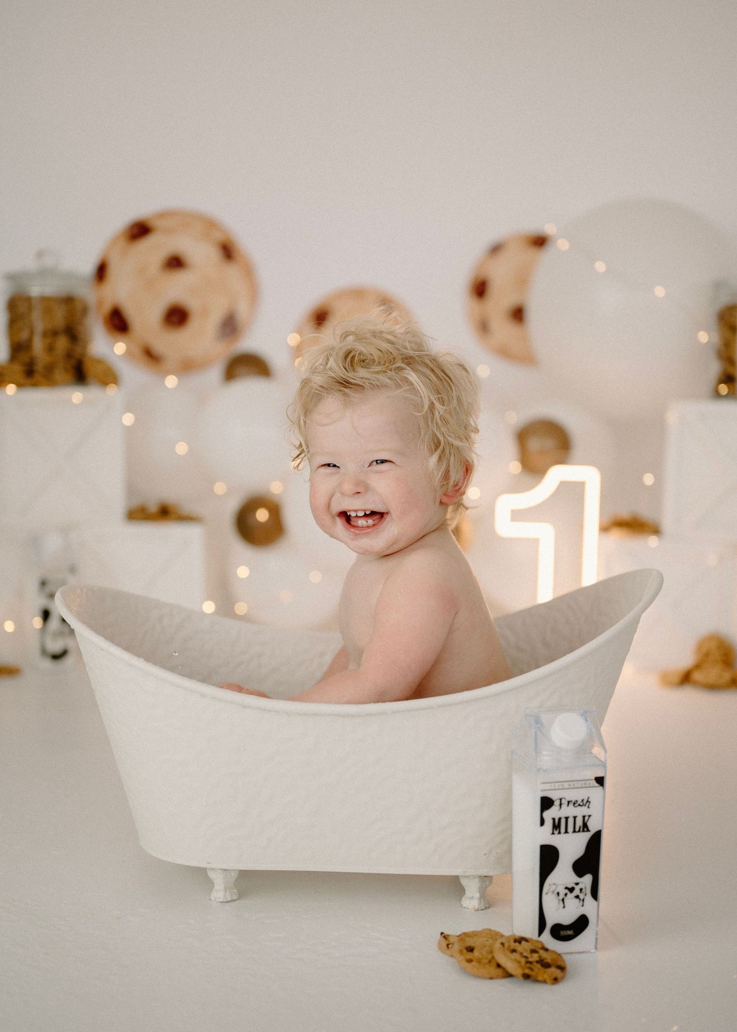 Laughing baby in white clawfoot tub at cookies and milk first birthday photo shoot with number 1 sign.