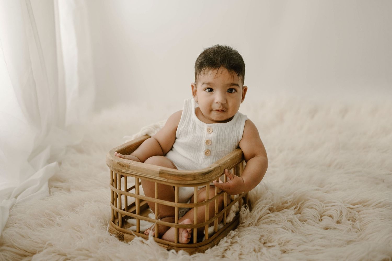 Cute baby in a white romper sitting inside a wicker basket on a fluffy cream rug in a bright studio.