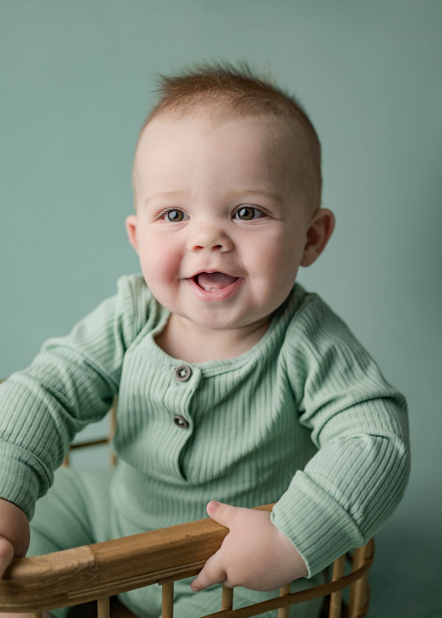 Laughing baby boy in green ribbed romper sitting in a wicker basket against a teal background.