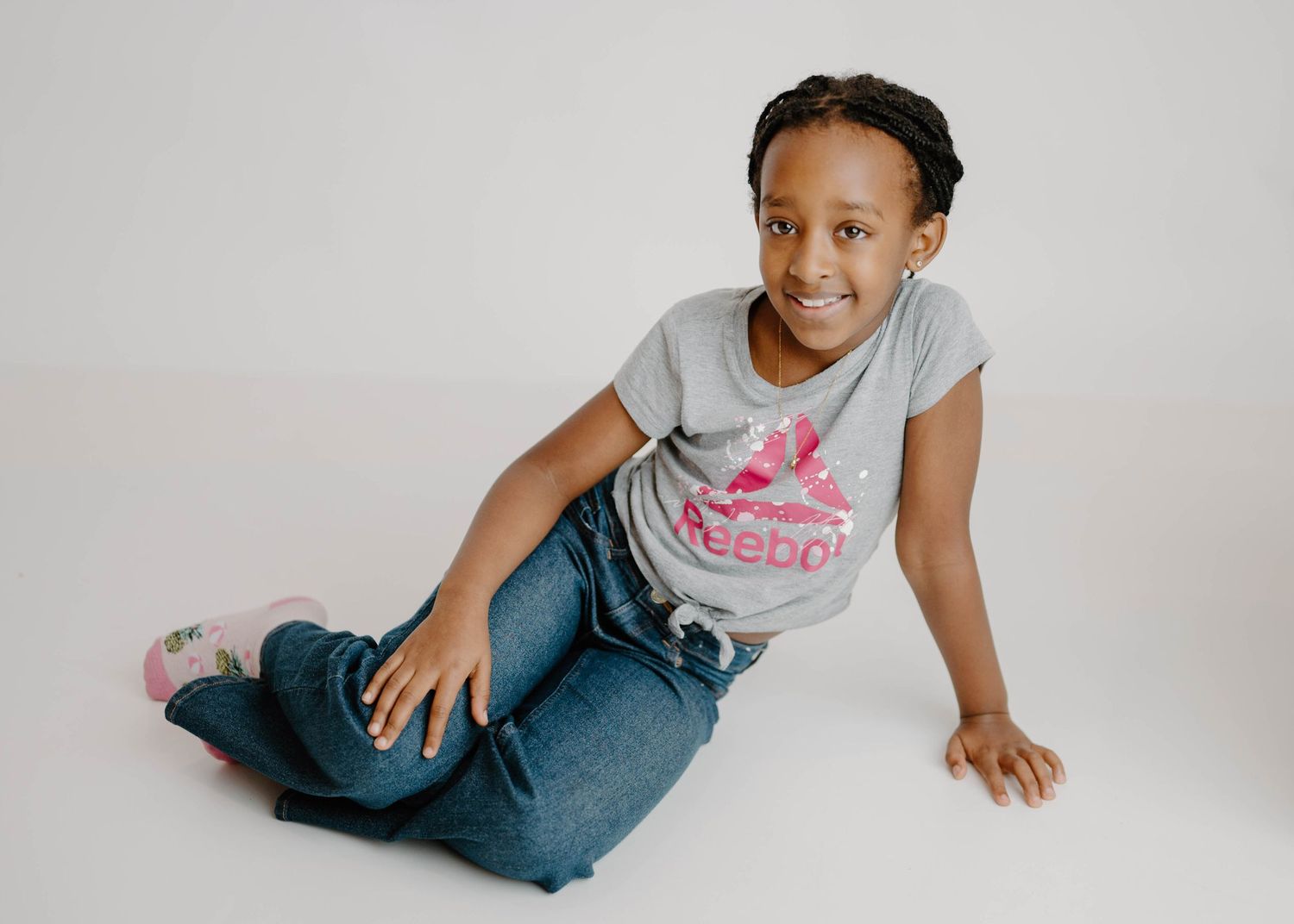Smiling young girl in gray Reebok tee and jeans sitting casually on white studio floor.