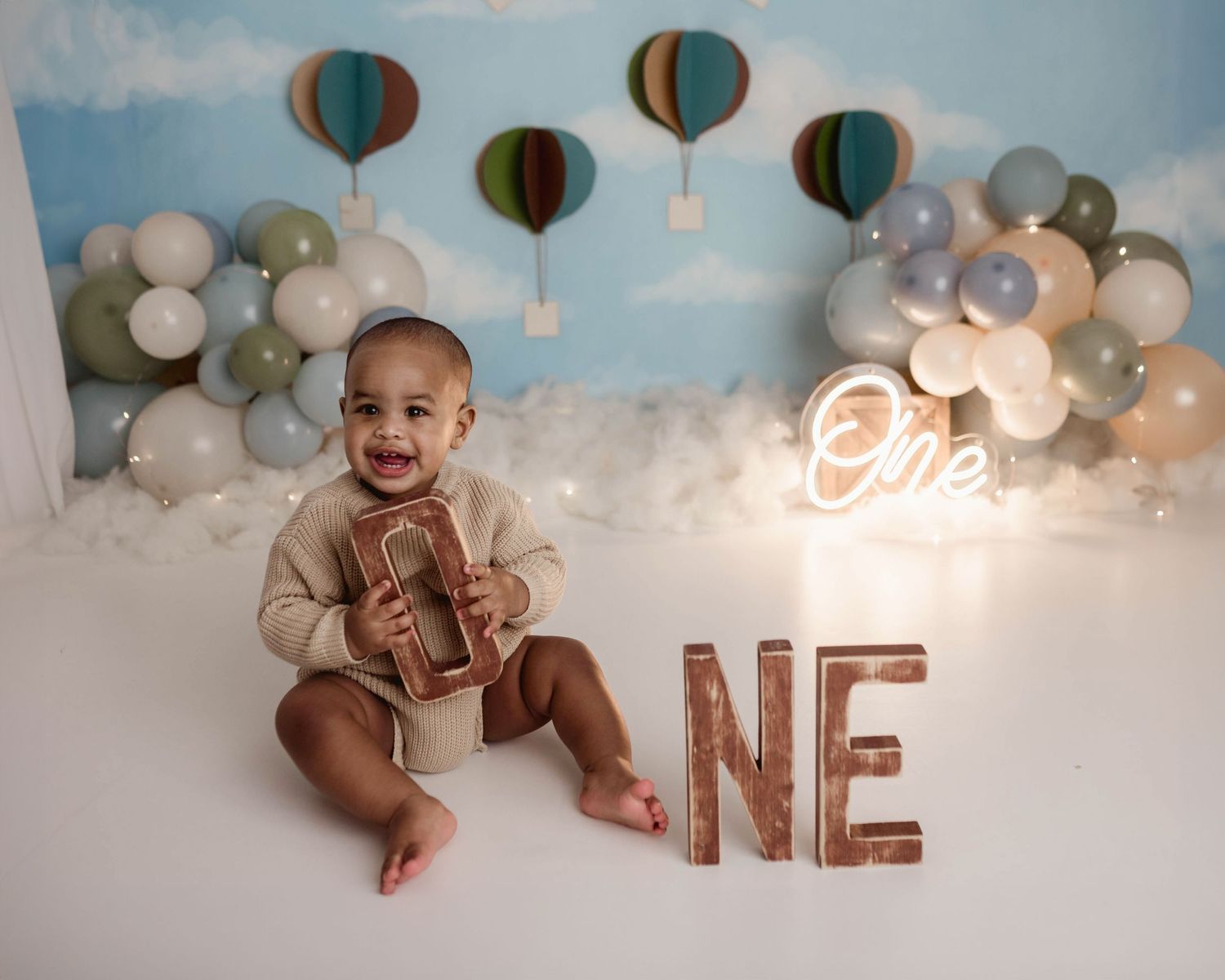 Happy baby holding wooden letter at a hot air balloon first birthday session with neon One sign and balloon garland.