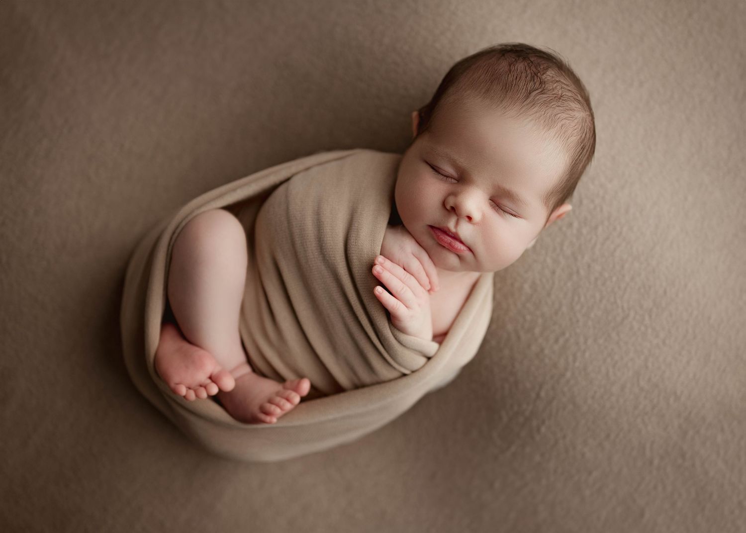 Sleeping newborn baby swaddled in beige wrap on a tan background.
