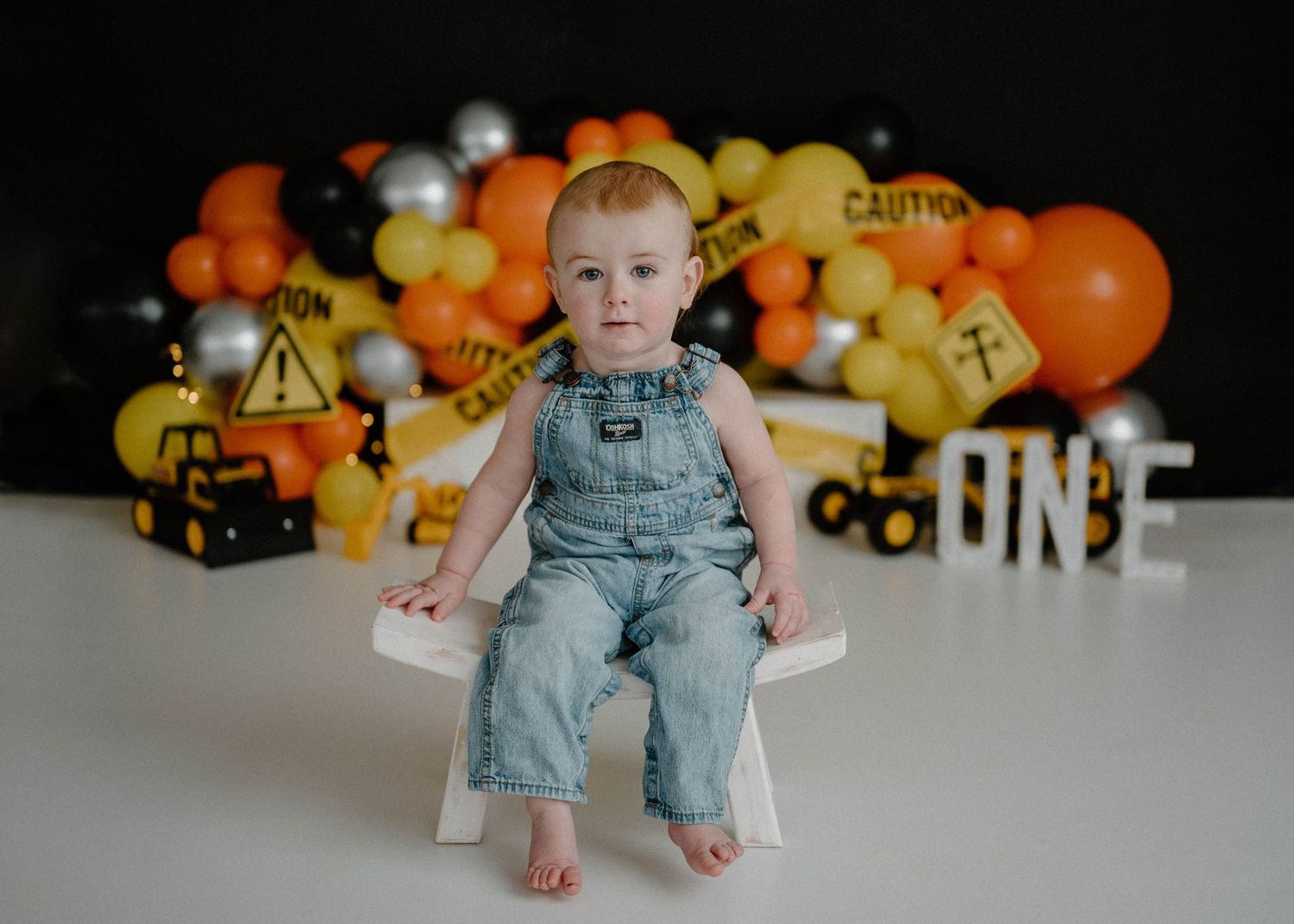 Baby boy in denim overalls poses before construction-themed ONE birthday backdrop with orange black balloons.