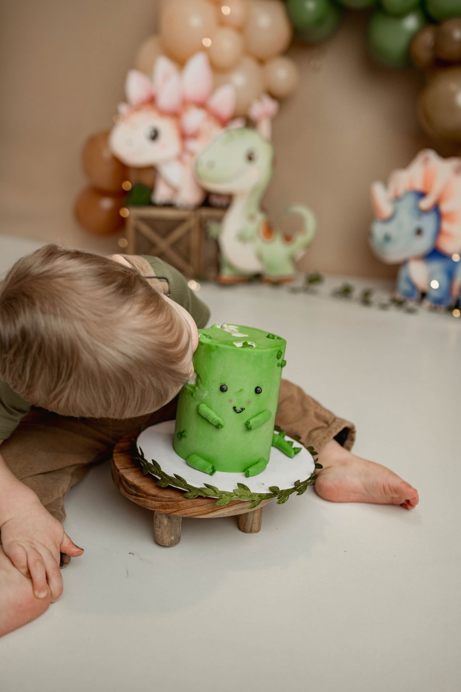 Baby boy bends down to smell a green dinosaur birthday cake surrounded by colorful dino decorations.
