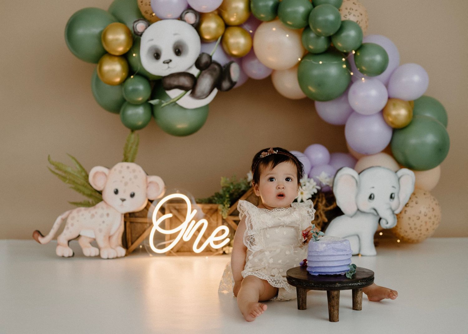 Baby girl in lace dress at jungle animal themed first birthday with panda and elephant decor.