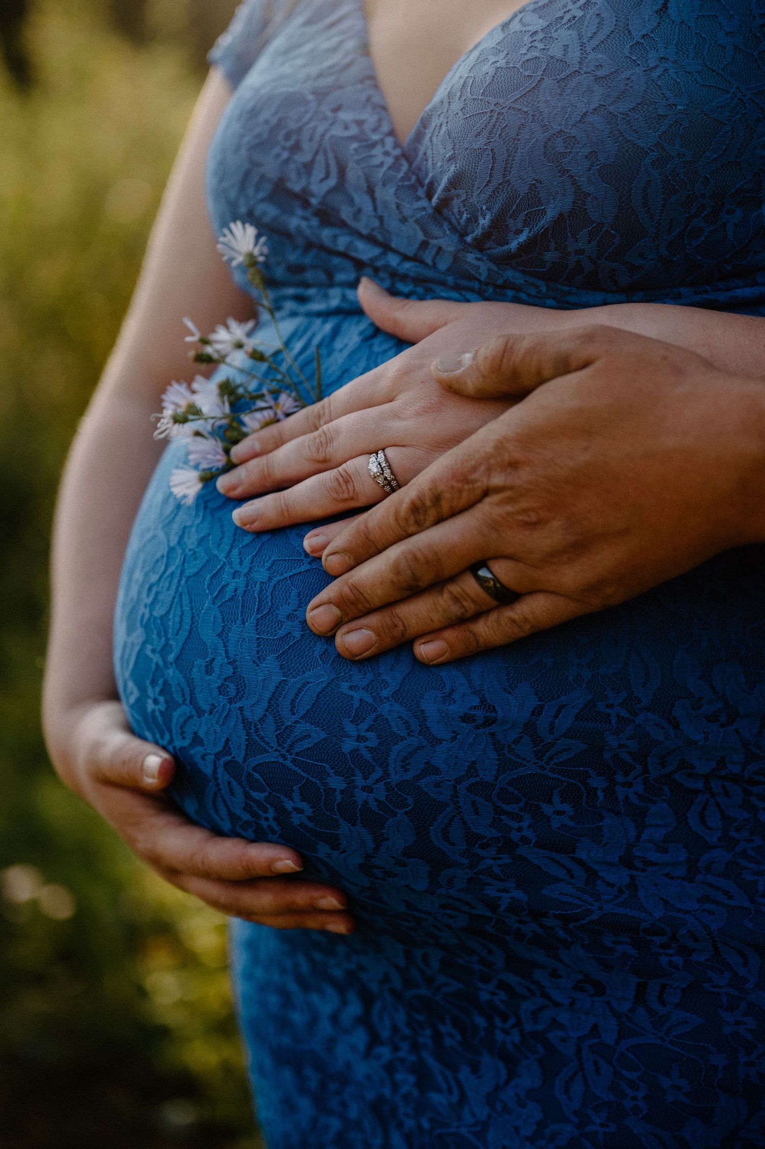 Close-up of pregnant couple holding baby bump with wildflowers in blue lace dress.
