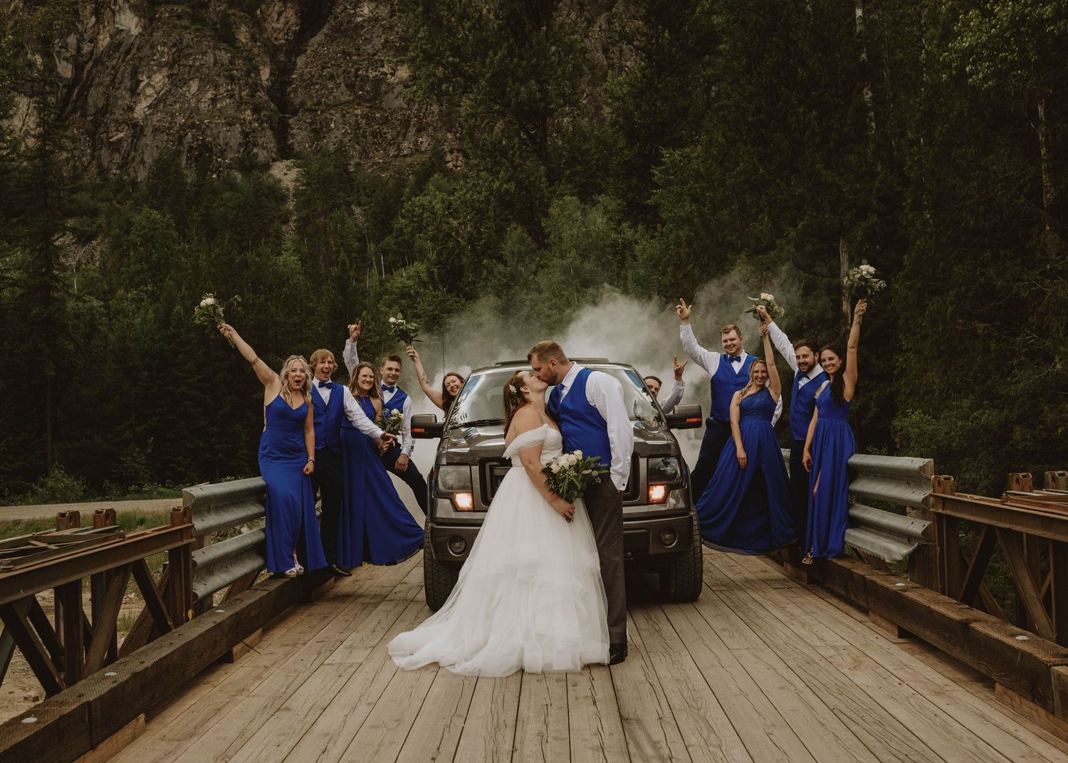 Wedding party in royal blue dresses celebrates with bride and groom in front of a black truck on a bridge.