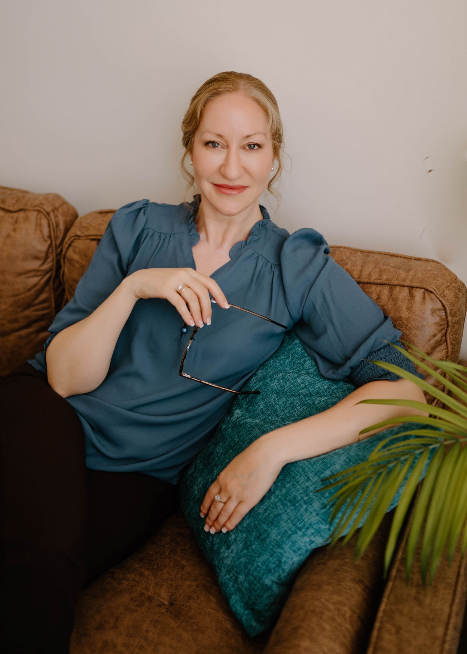 Elegant blonde woman in teal blouse relaxing on brown leather couch holding glasses, smiling.