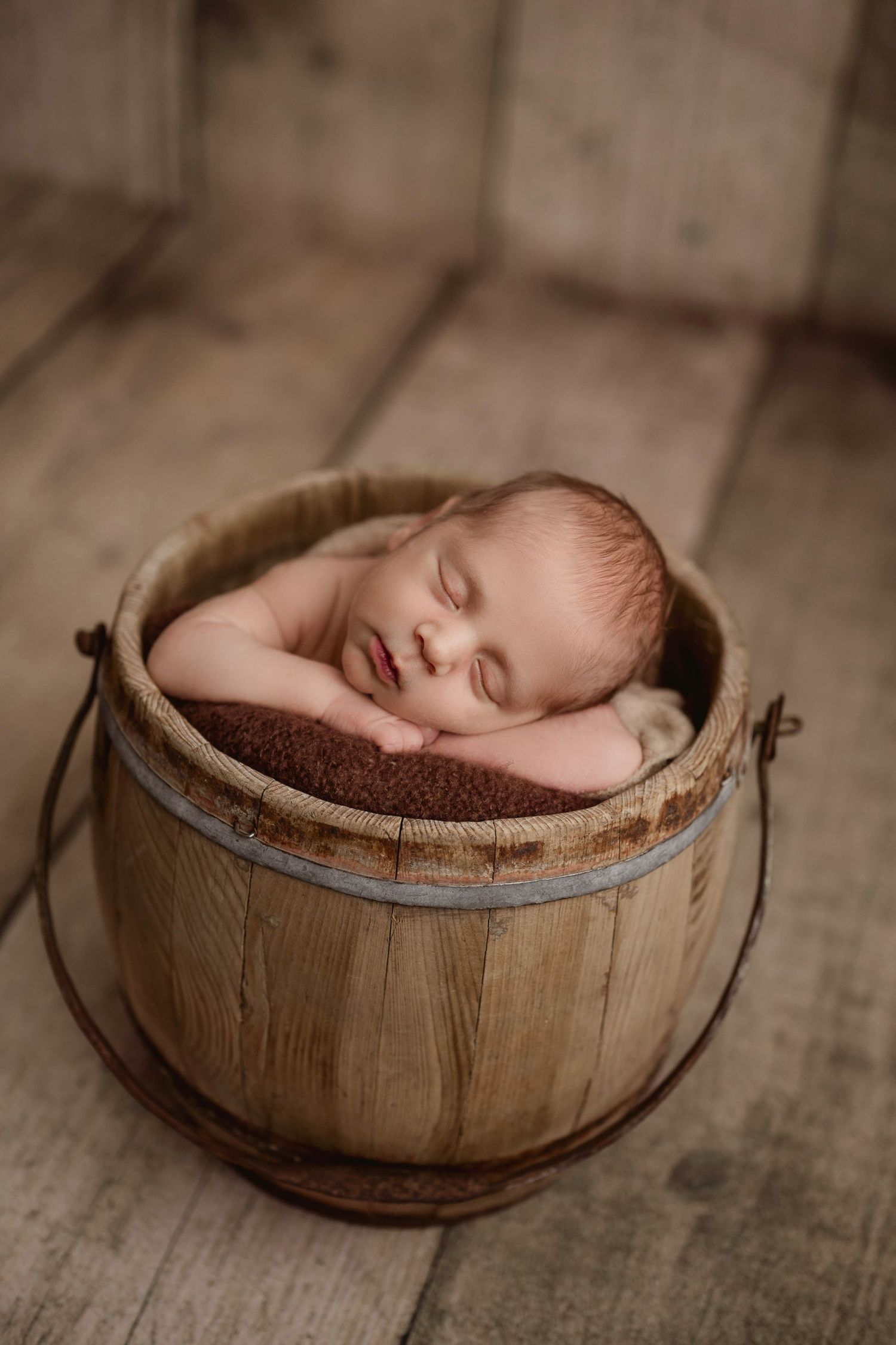 Peaceful sleeping newborn baby resting in rustic wooden bucket with brown blanket on wood floor.