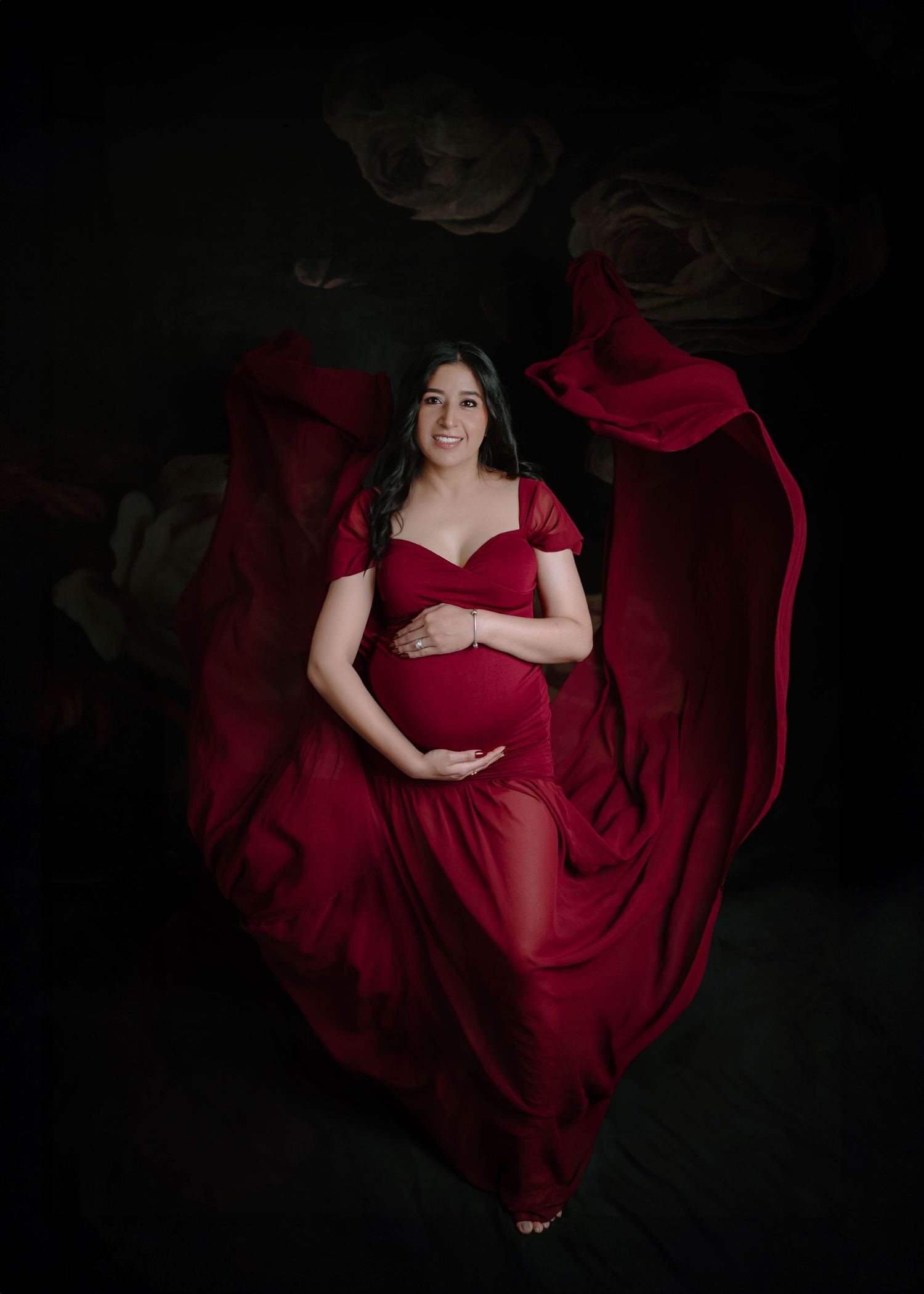 A pregnant woman in a dramatic red flowing gown poses with fabric spread wide in a dark floral studio.
