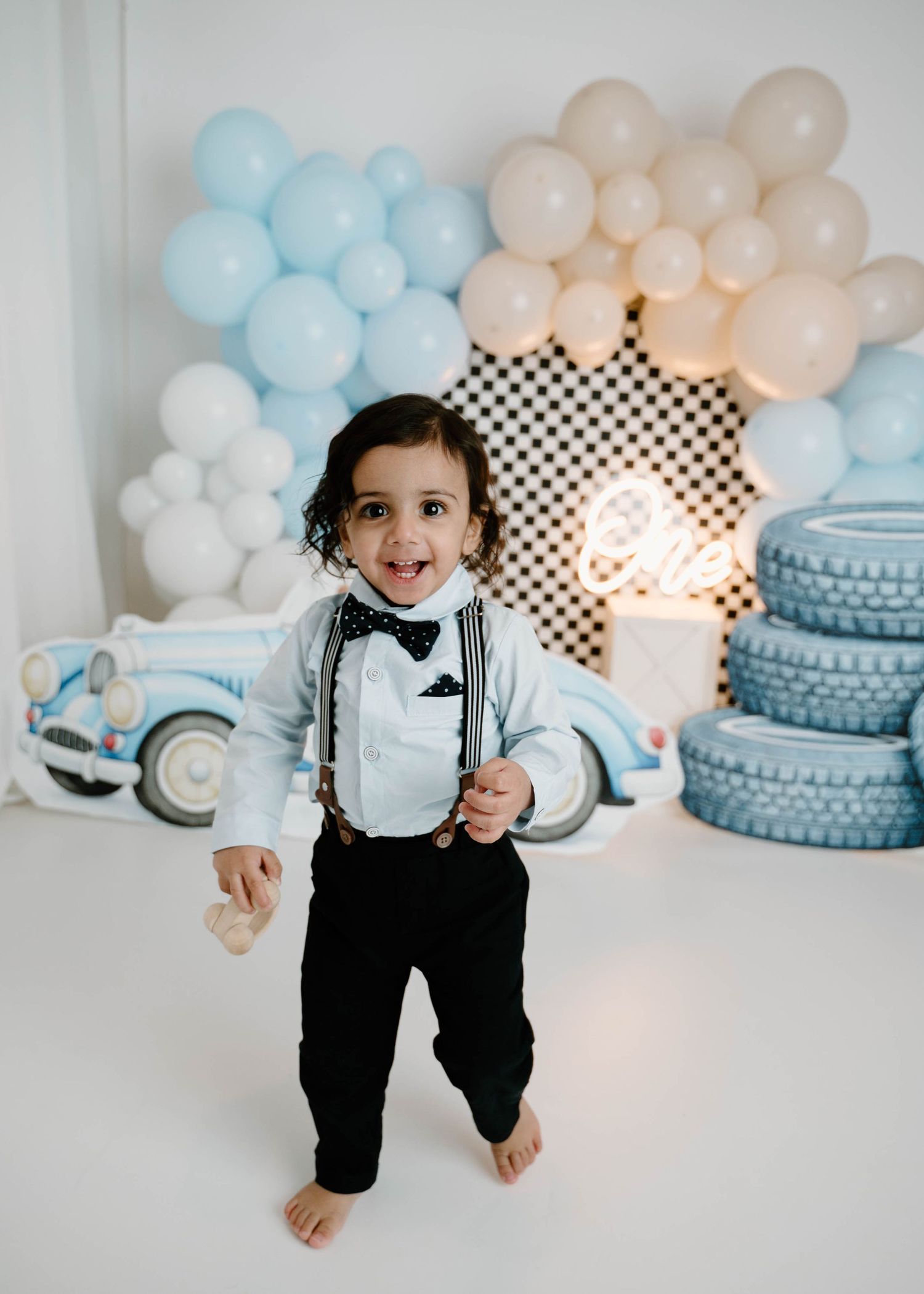 A happy toddler boy in suspenders and bow tie stands barefoot in front of a blue vintage race car balloon backdrop.