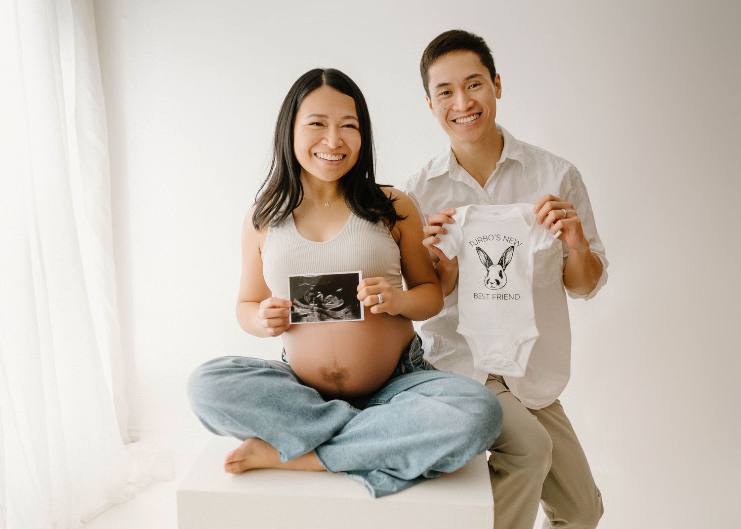 Smiling couple holding ultrasound photo and baby onesie during maternity photo session.