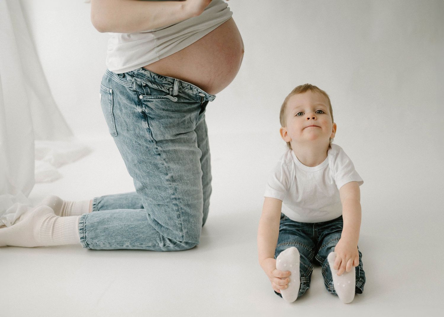 Pregnant woman kneels beside seated toddler boy looking up in a minimalist white studio.