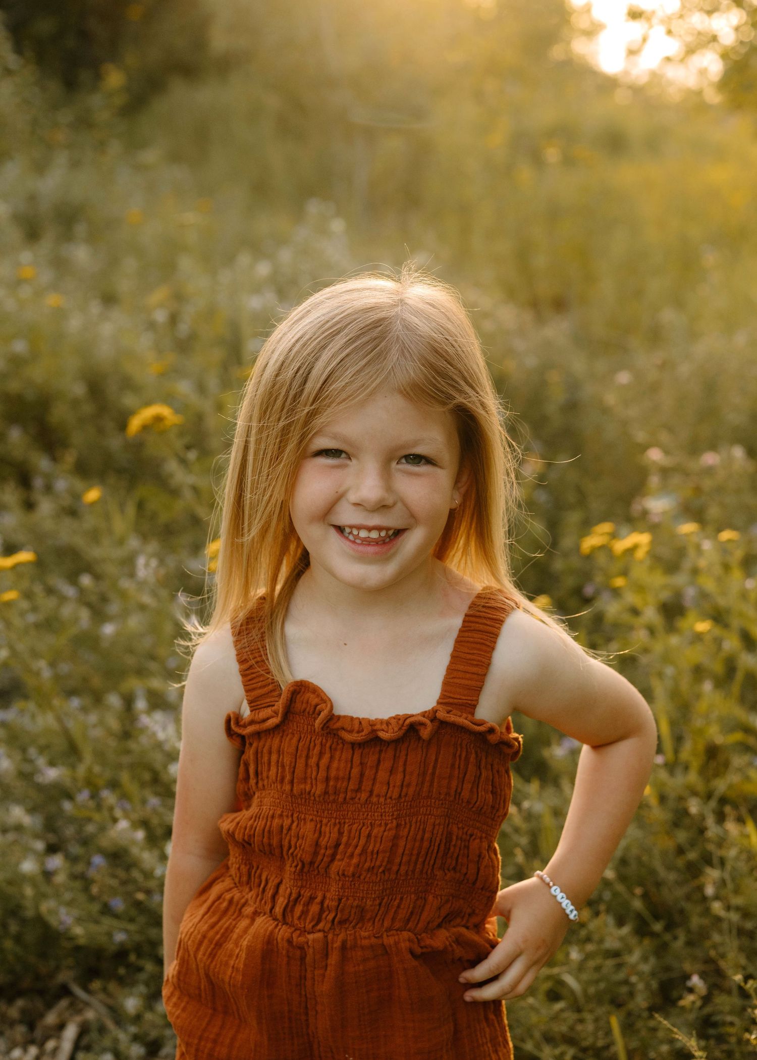 Smiling young girl in rust orange romper posing in golden wildflower field at sunset.