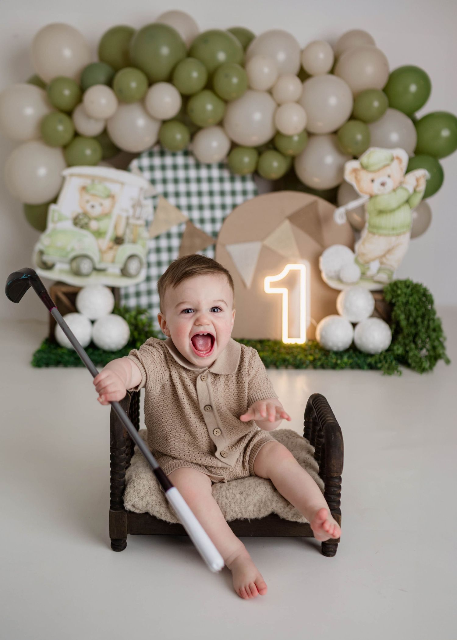 Laughing baby boy sitting in chair holding golf club at golf-themed first birthday party with balloon arch.
