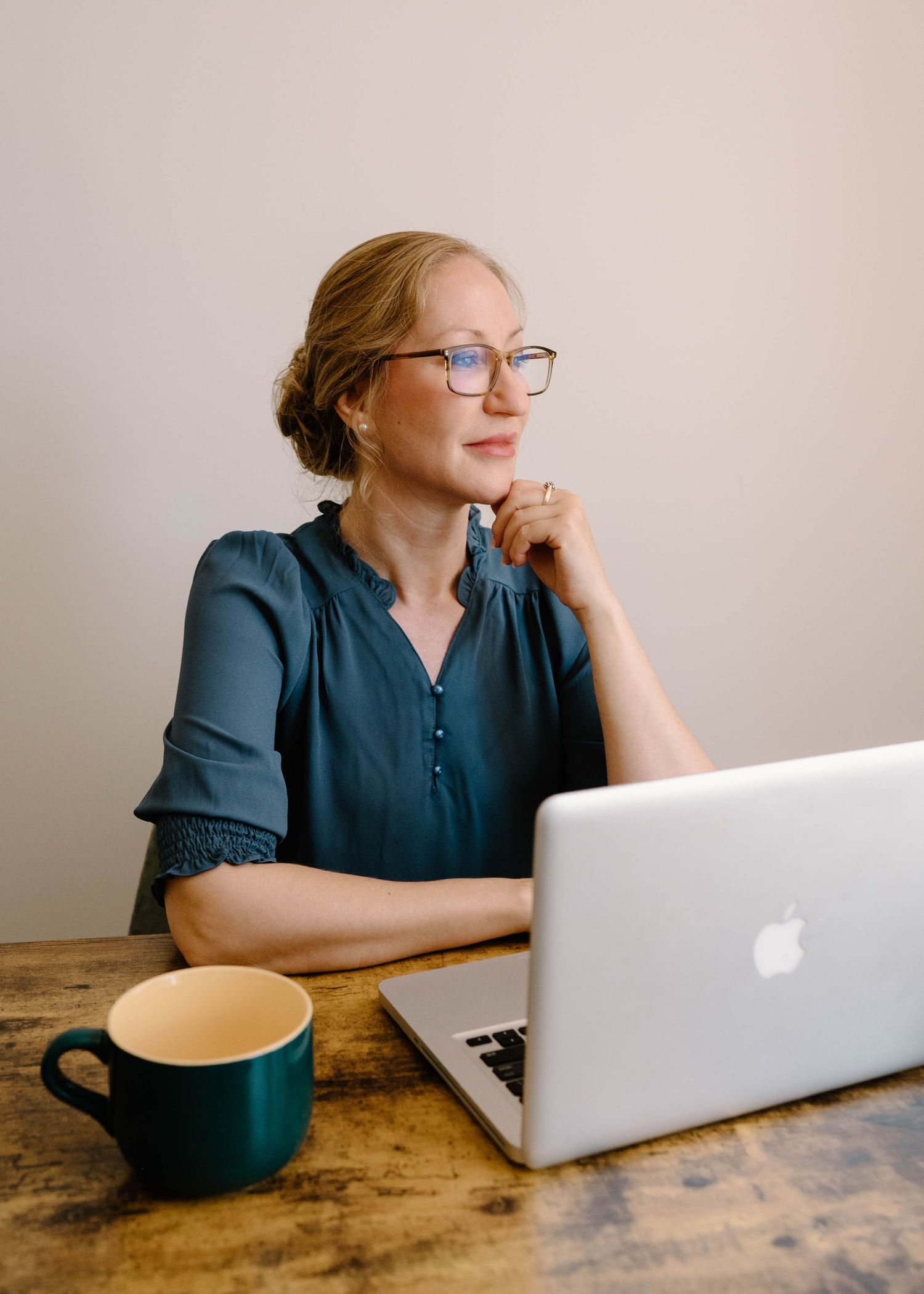 Professional woman with glasses working on Apple MacBook laptop at wooden desk with coffee mug.
