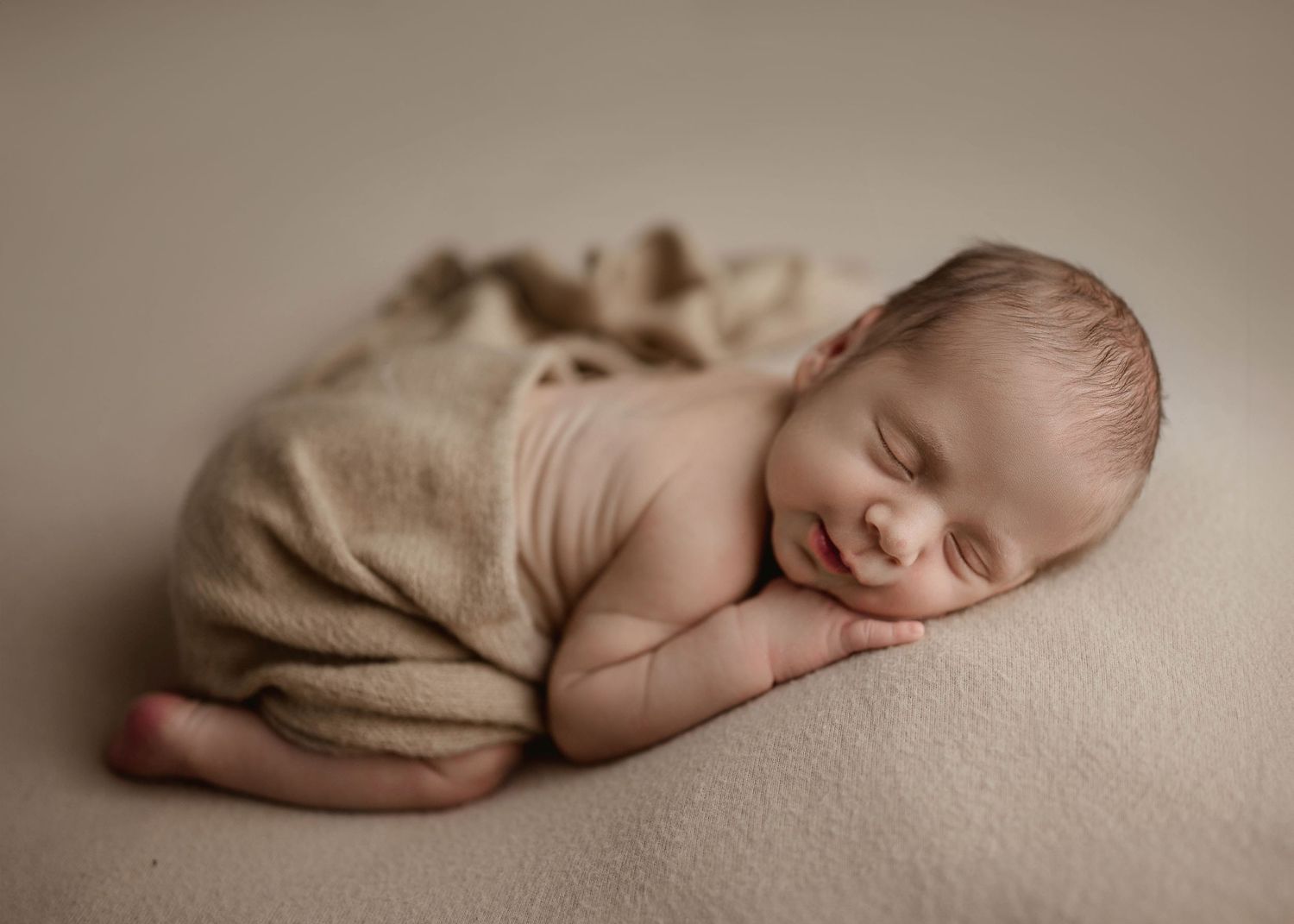 Smiling newborn baby wrapped in beige knit blanket sleeping peacefully on soft cream backdrop.